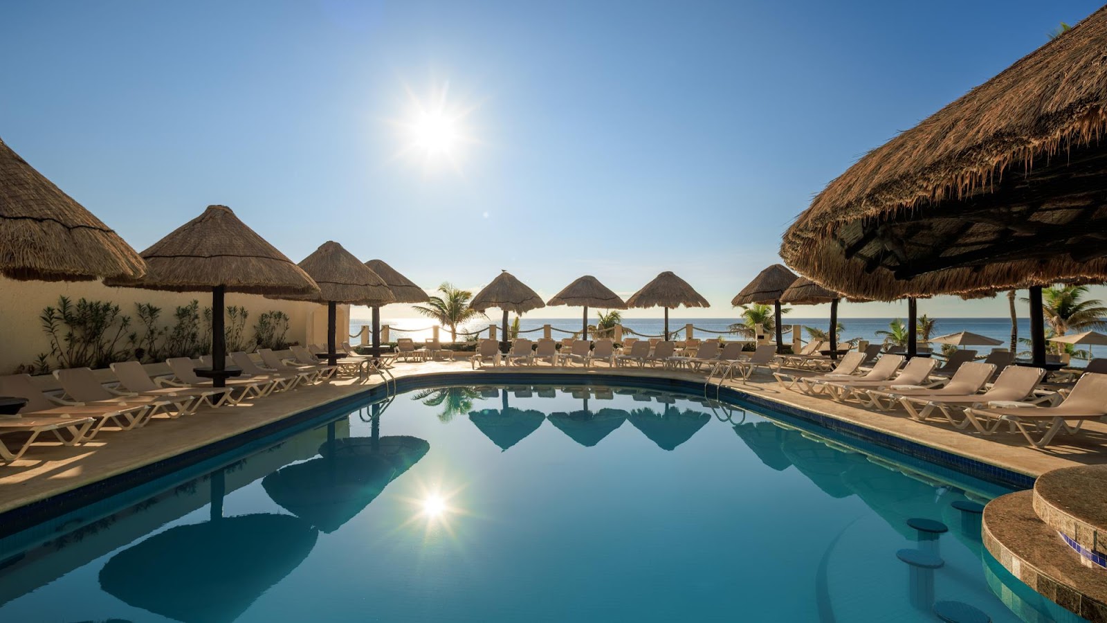 Resort pool with thatched palapas, loungers, and ocean view under clear blue sky at Occidental Tucancún.