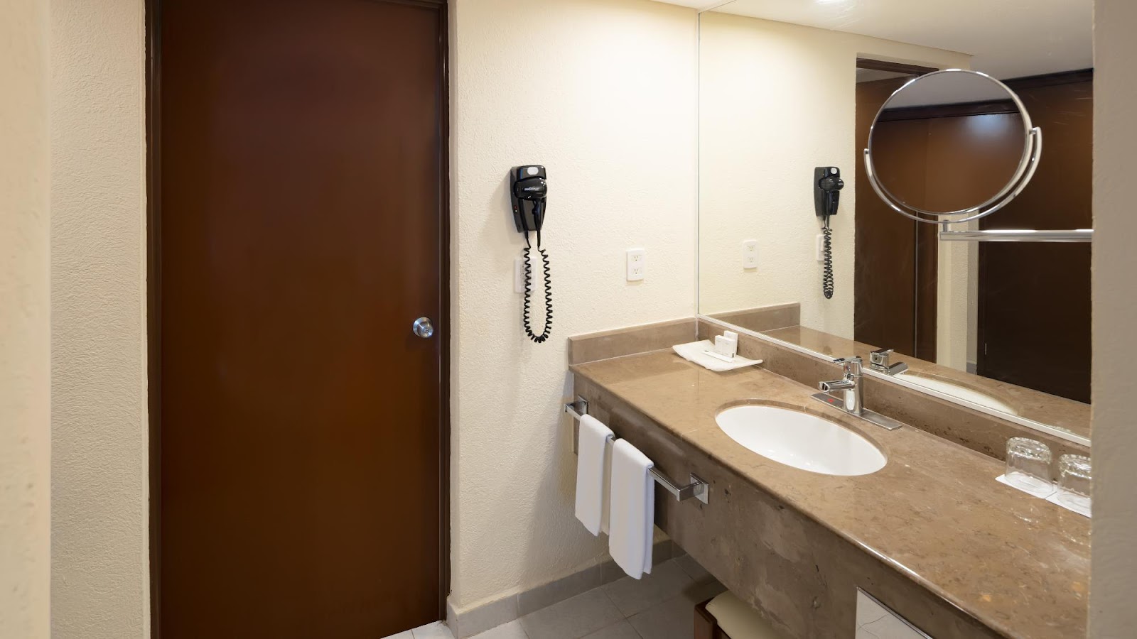 Bathroom vanity with tan countertop, white oval sink, large mirror, and two wall-mounted phones at Occidental Tucancún.