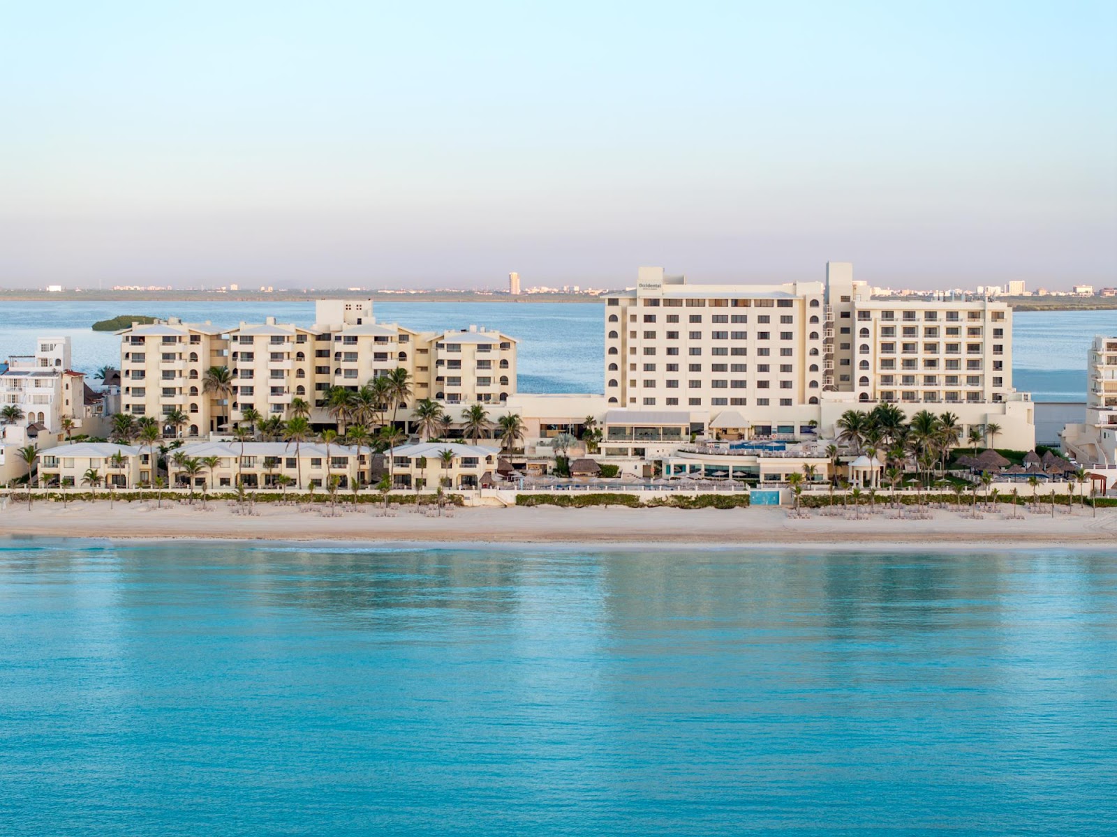Aerial view of Occidental Tucancún resort with white buildings, blue pools, and white loungers on a sandy beach beside turquoise water.
