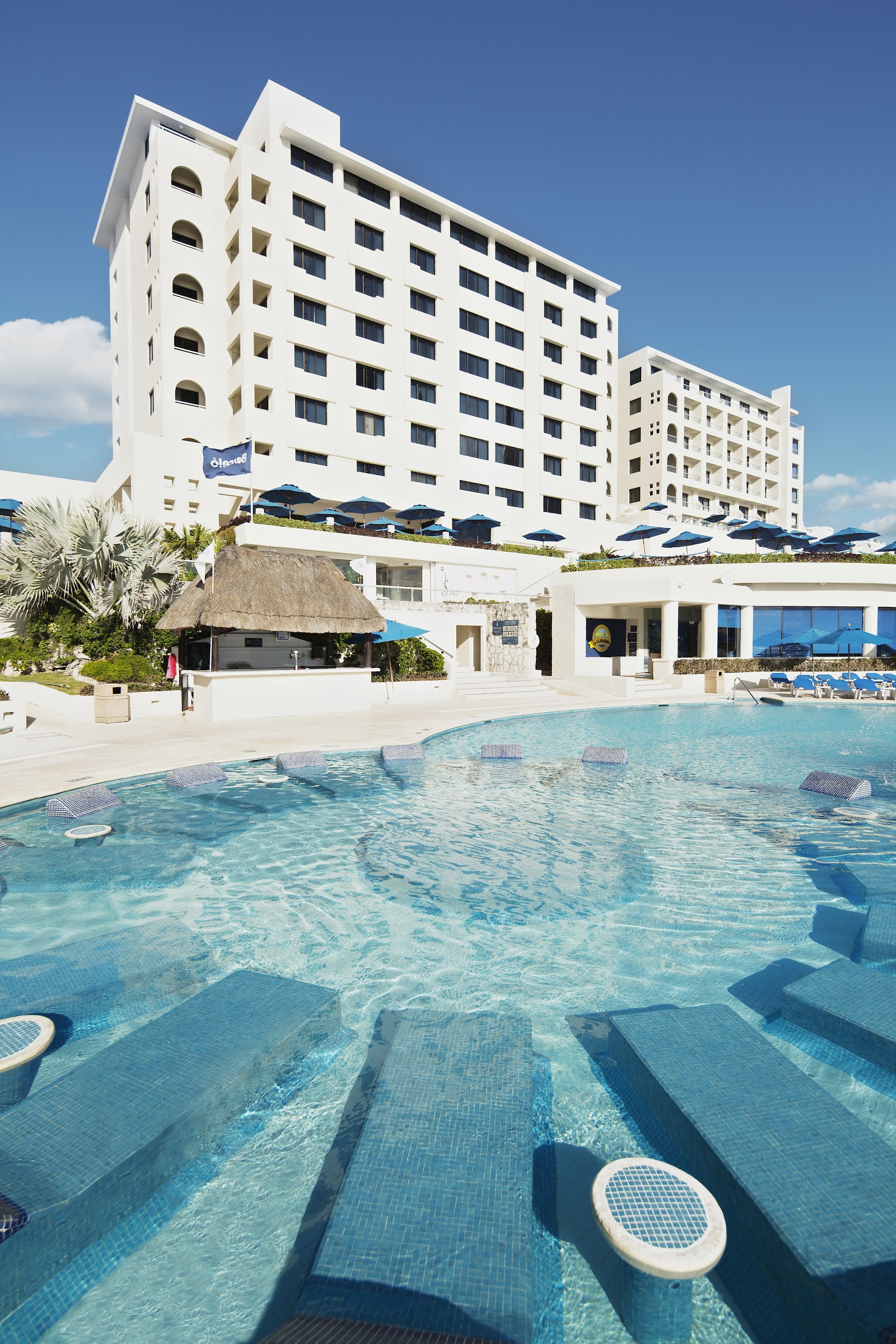 White multi-story resort buildings overlook a shallow pool with blue umbrellas, thatched-roof bar, and clear turquoise water under blue sky.