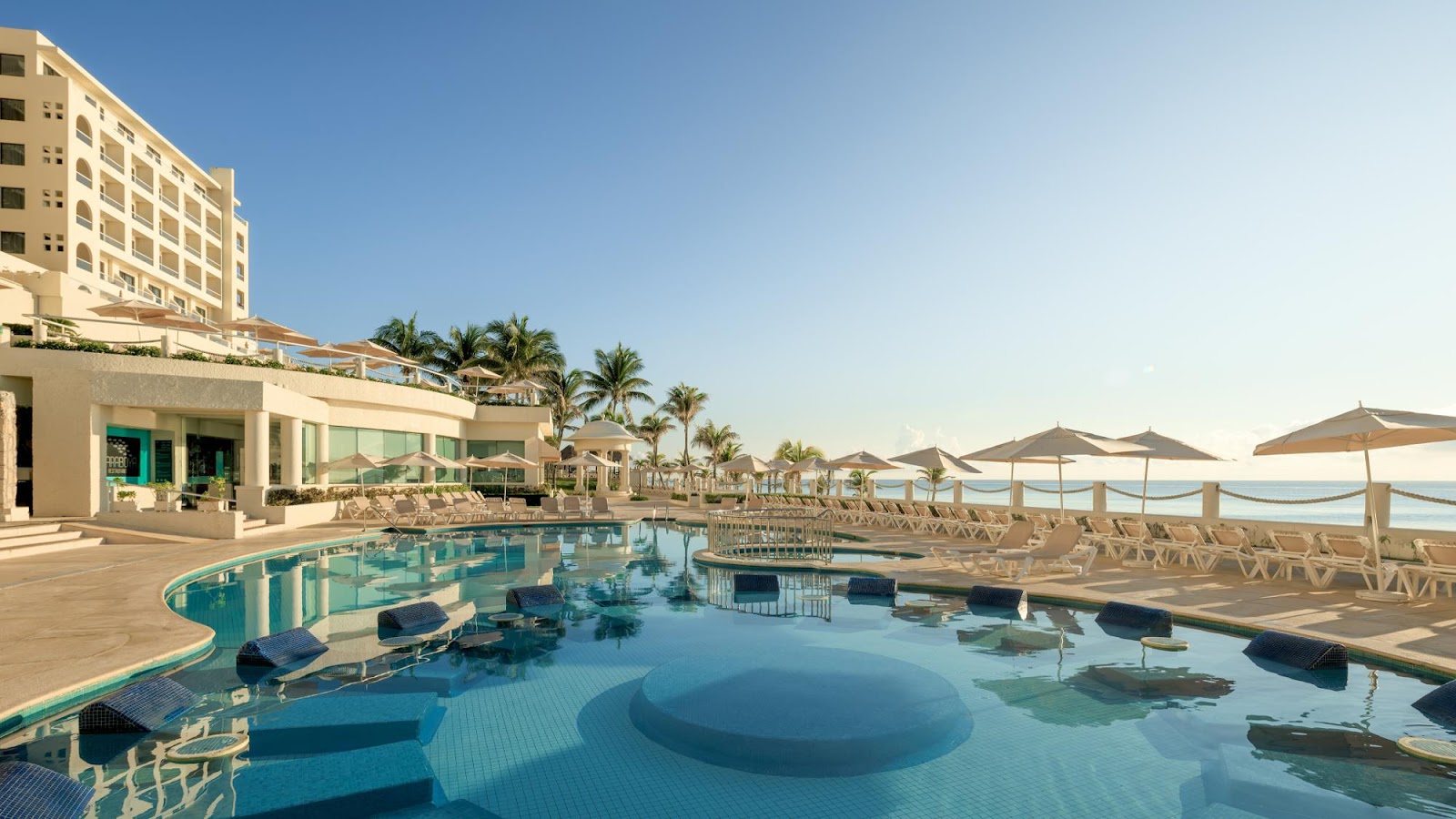 Beachfront pool with sunken lounge areas at Occidental Tucancún, flanked by palm trees, umbrellas, and a multi-story resort building.