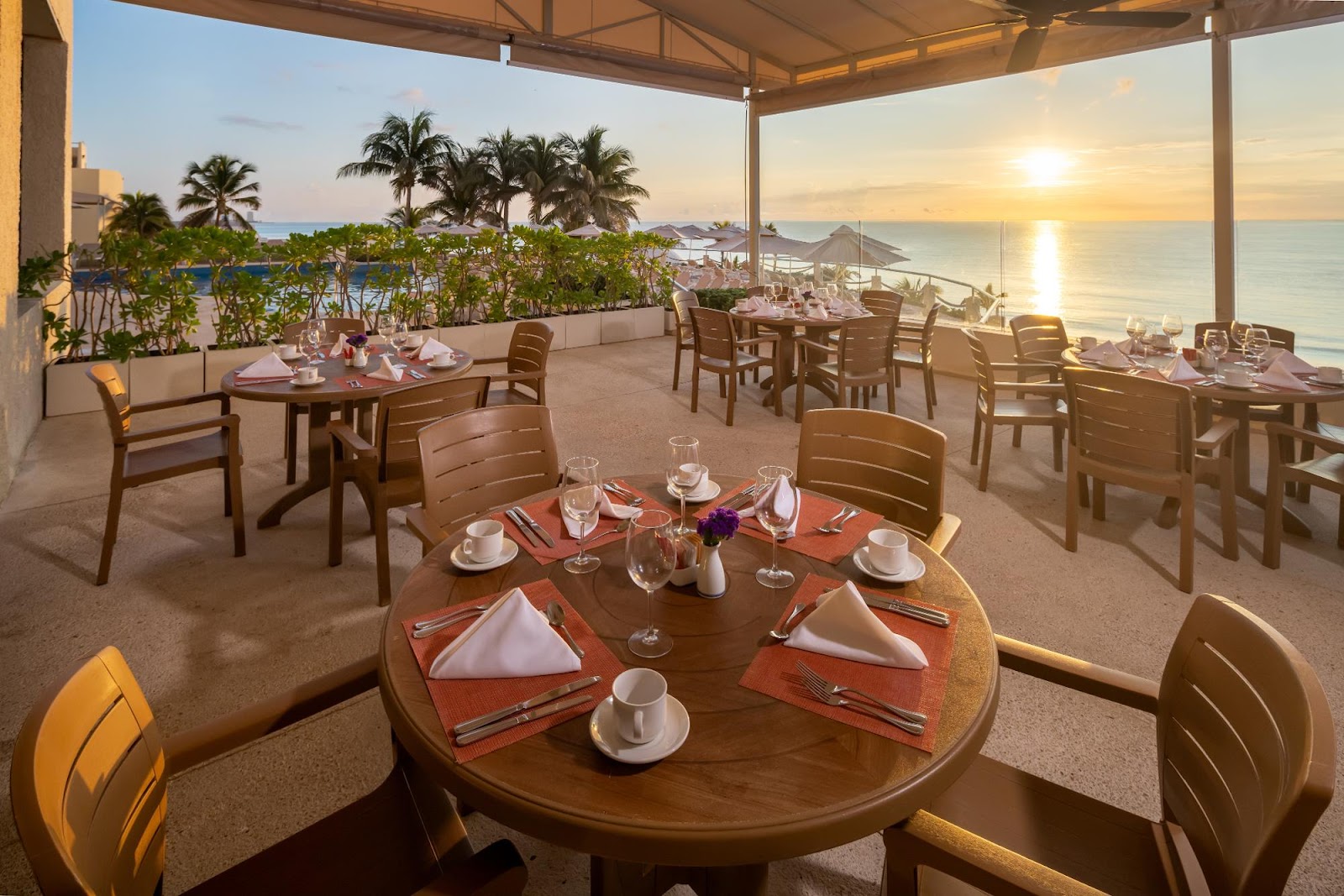 Covered dining terrace at sunset with round tables set for dinner, ocean and palm trees visible beyond, mountains in distance.