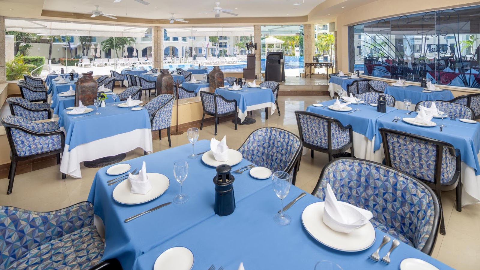 Dining room with blue tablecloths, patterned chairs, and place settings overlooking the pool and beach at Occidental Tucancún.