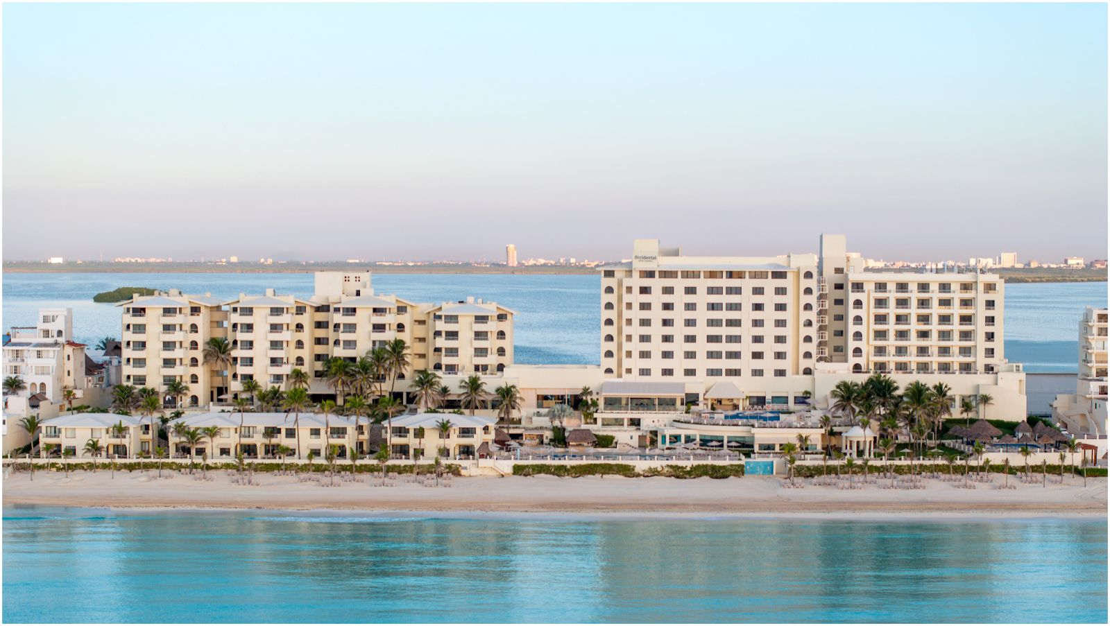 Tan and cream-colored resort buildings with palm trees facing a sandy beach and turquoise water at Occidental Tucancún.