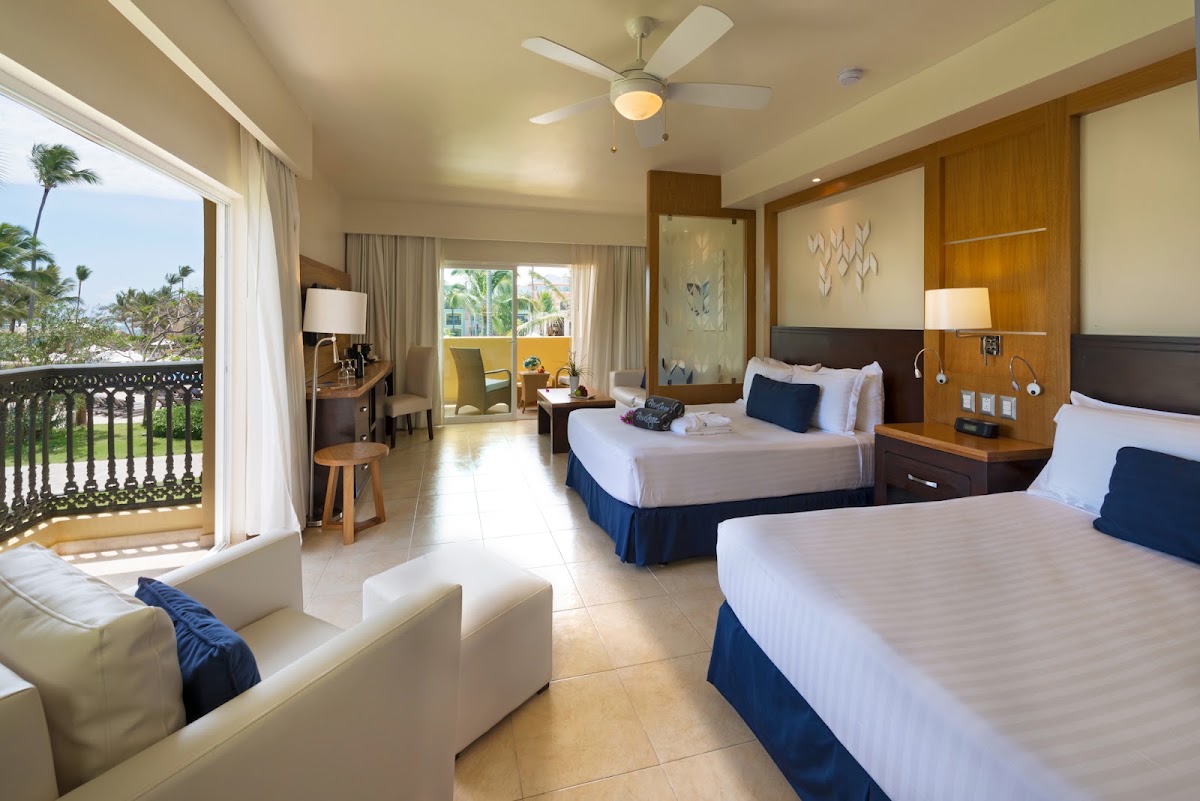 Spacious resort room with two double beds, wooden accents, and a balcony overlooking palm trees and ocean grounds.