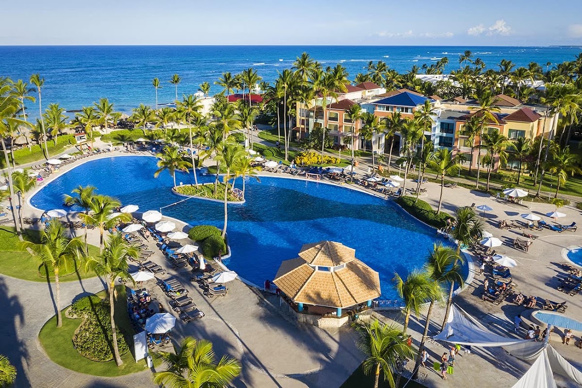 Large resort pool with thatched-roof bar surrounded by palm trees, loungers, and beachfront buildings overlooking blue ocean water.
