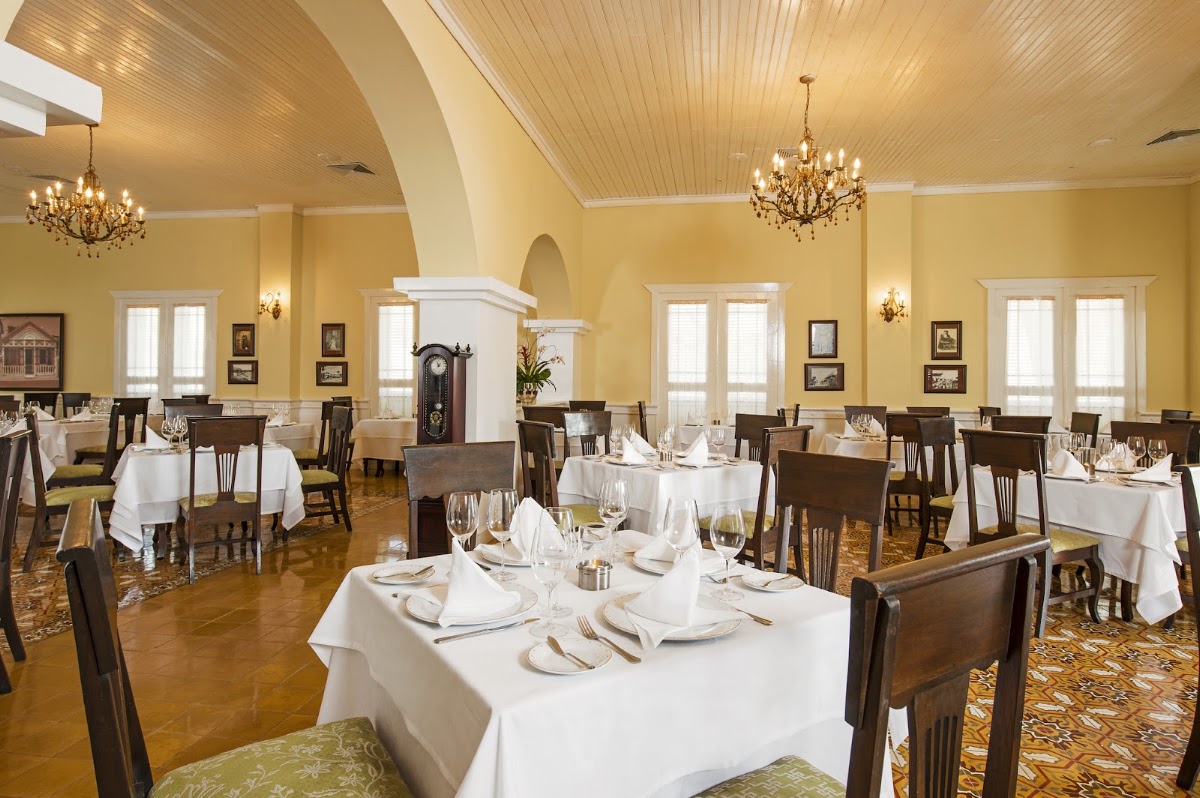 Elegant dining room with round tables set with white linens, dark wood chairs, crystal chandeliers, and yellow walls with arched ceiling.