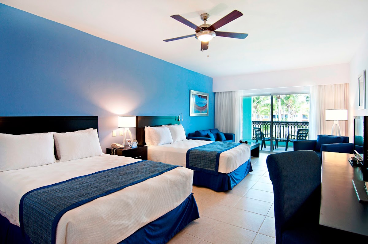 Resort hotel room with two double beds, blue accent wall, ceiling fan, and ocean view balcony with palm trees visible through glass doors.