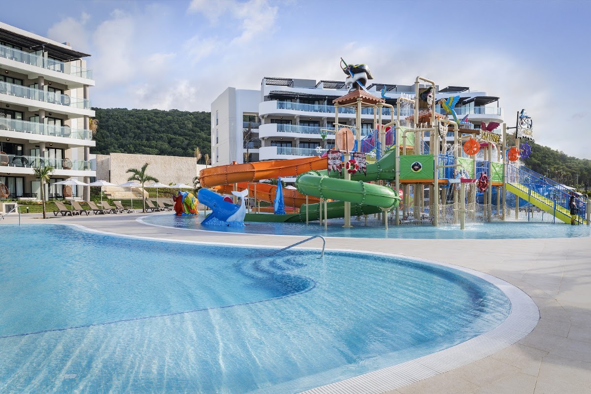 Curved pool in foreground with colorful water slides, splash pad, and multi-story play structure at Ocean Coral Spring, forested hillside behind modern resort buildings.