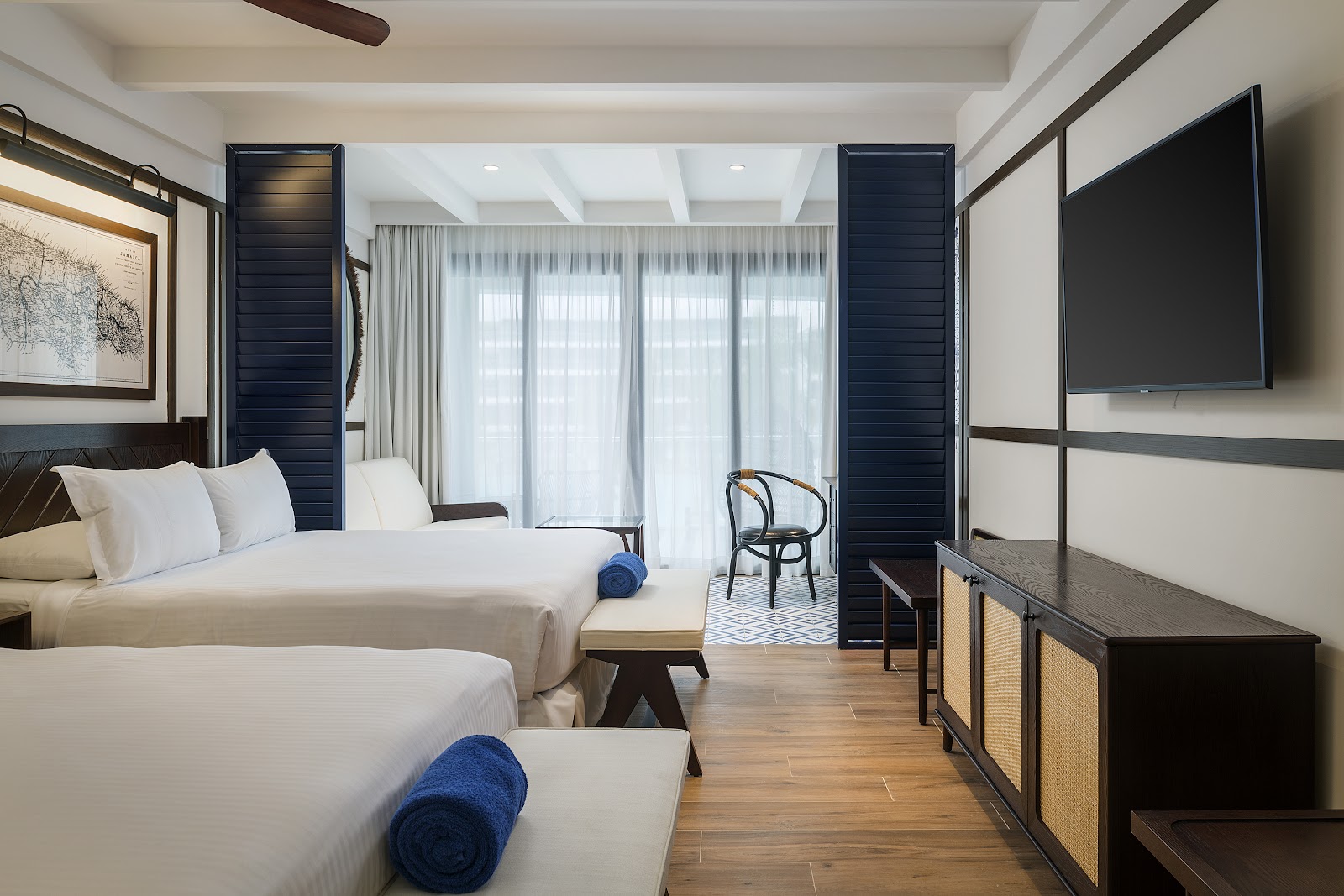 Modern resort bedroom with two queen beds, blue accent pillows, dark wood headboards, and sheer curtains framing a balcony door.