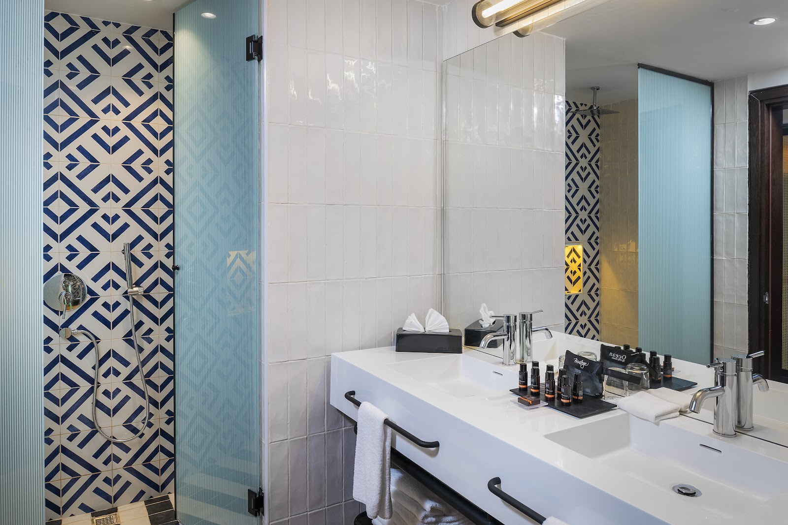 Modern bathroom with blue and cream geometric tile accent wall, white subway tiles, glass shower enclosure, and white vanity with amenities and towels.