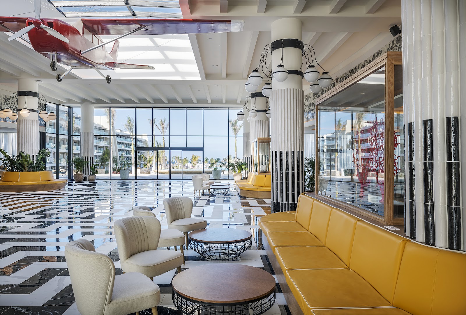 Modern lobby with geometric floor patterns, cream and yellow seating, cylindrical pendant lights, and ocean views through floor-to-ceiling windows at Ocean Coral Spring.