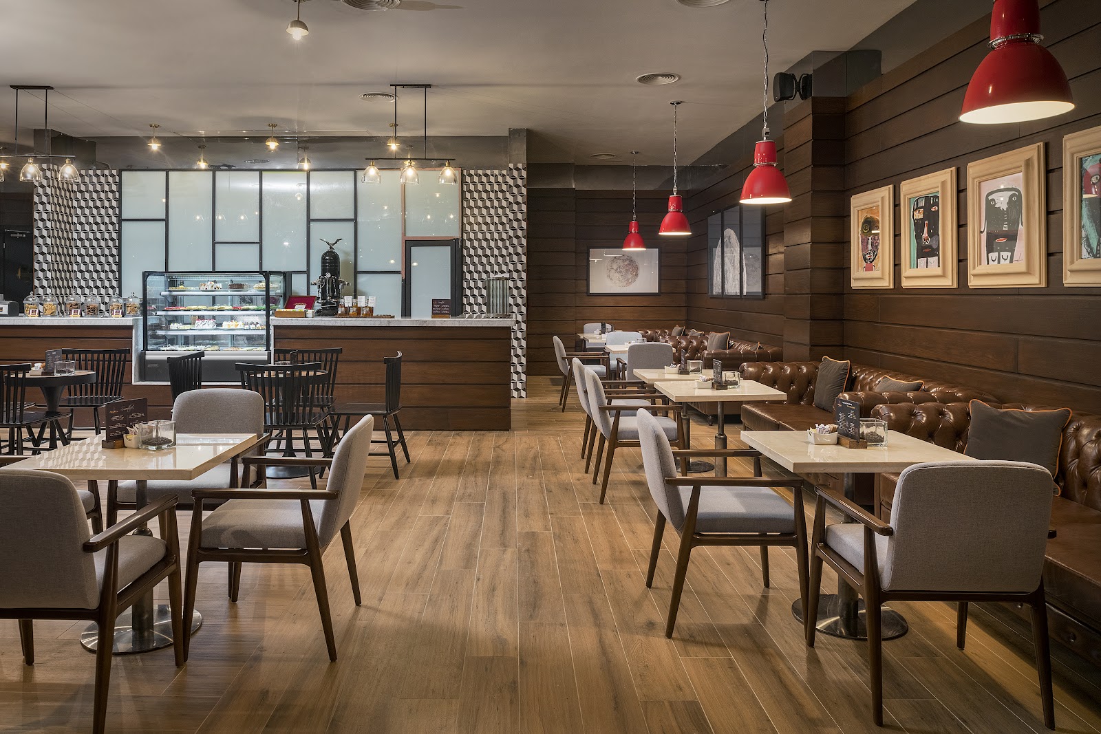 Modern cafe interior with wood paneling, dining tables, leather booth seating, red pendant lights, and a pastry counter with glass display case.