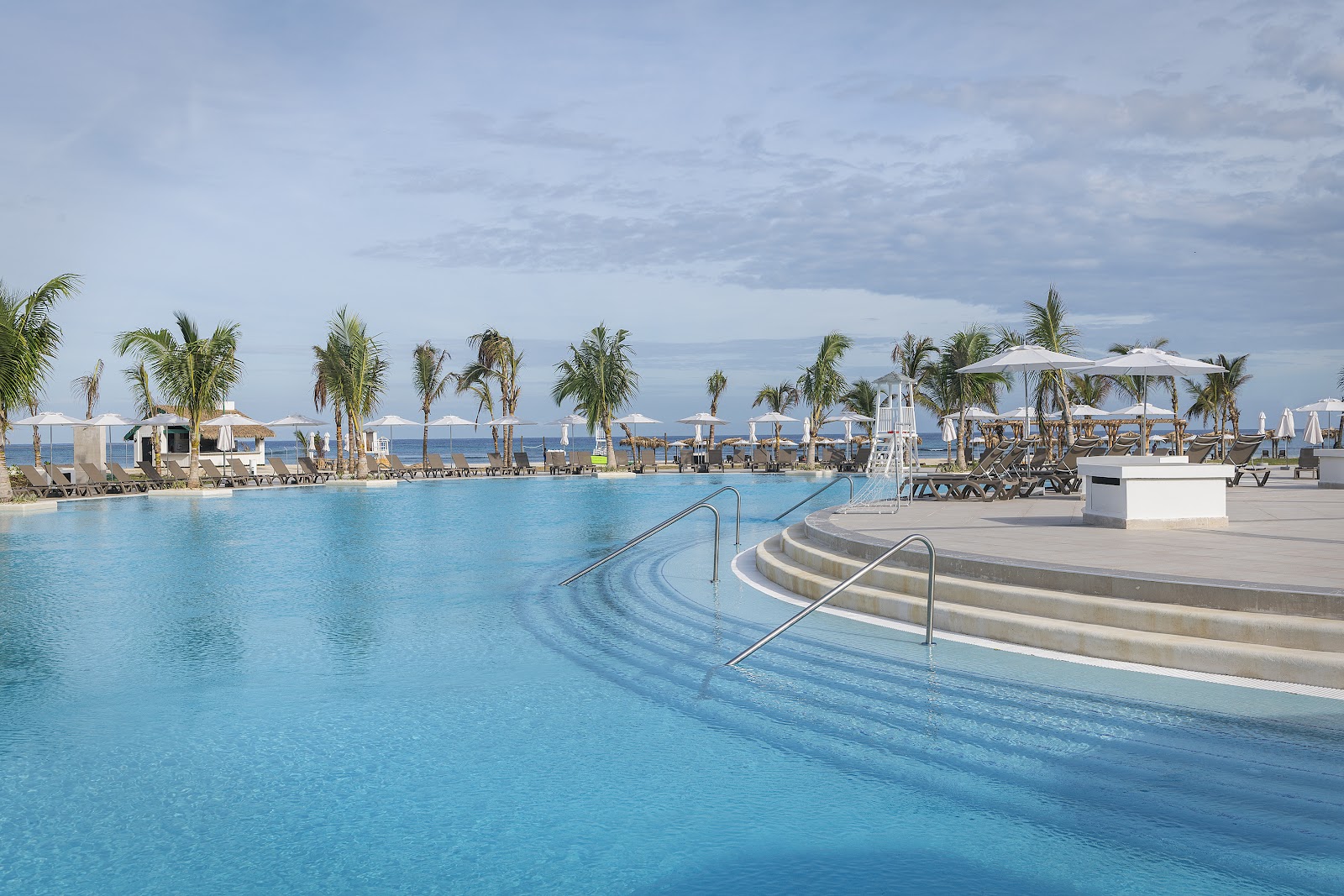 Large lagoon-style pool with stepped entry and metal railings, bordered by palm trees, white umbrellas, lounge chairs, and the ocean beyond under cloudy sky.