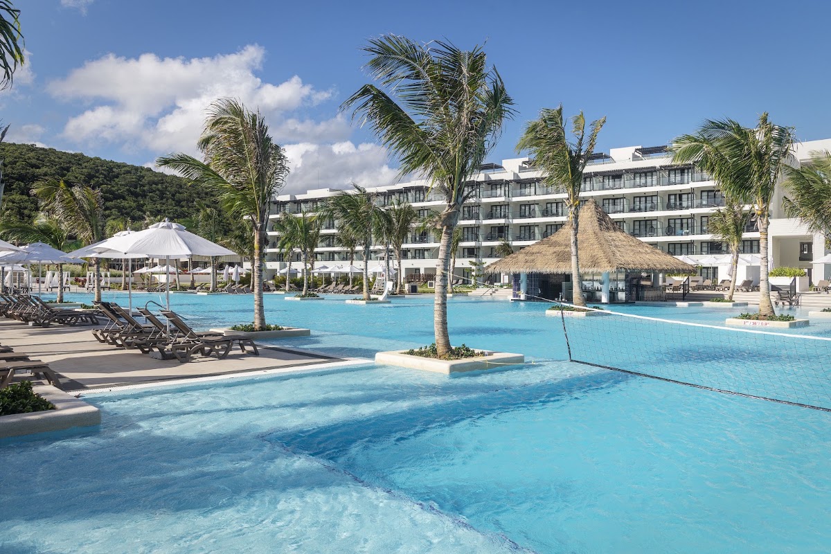 Multi-level turquoise pool with palm trees, loungers, umbrellas, and thatched bar structure at Ocean Eden Bay resort, with white building and hillside beyond.