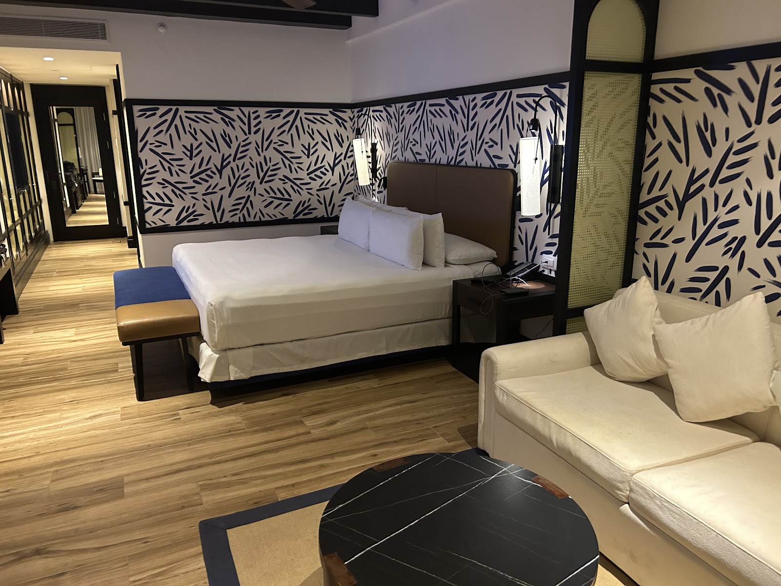 Resort suite with queen bed, cream seating area, black marble table, and leaf-patterned headboard wall on wood flooring.