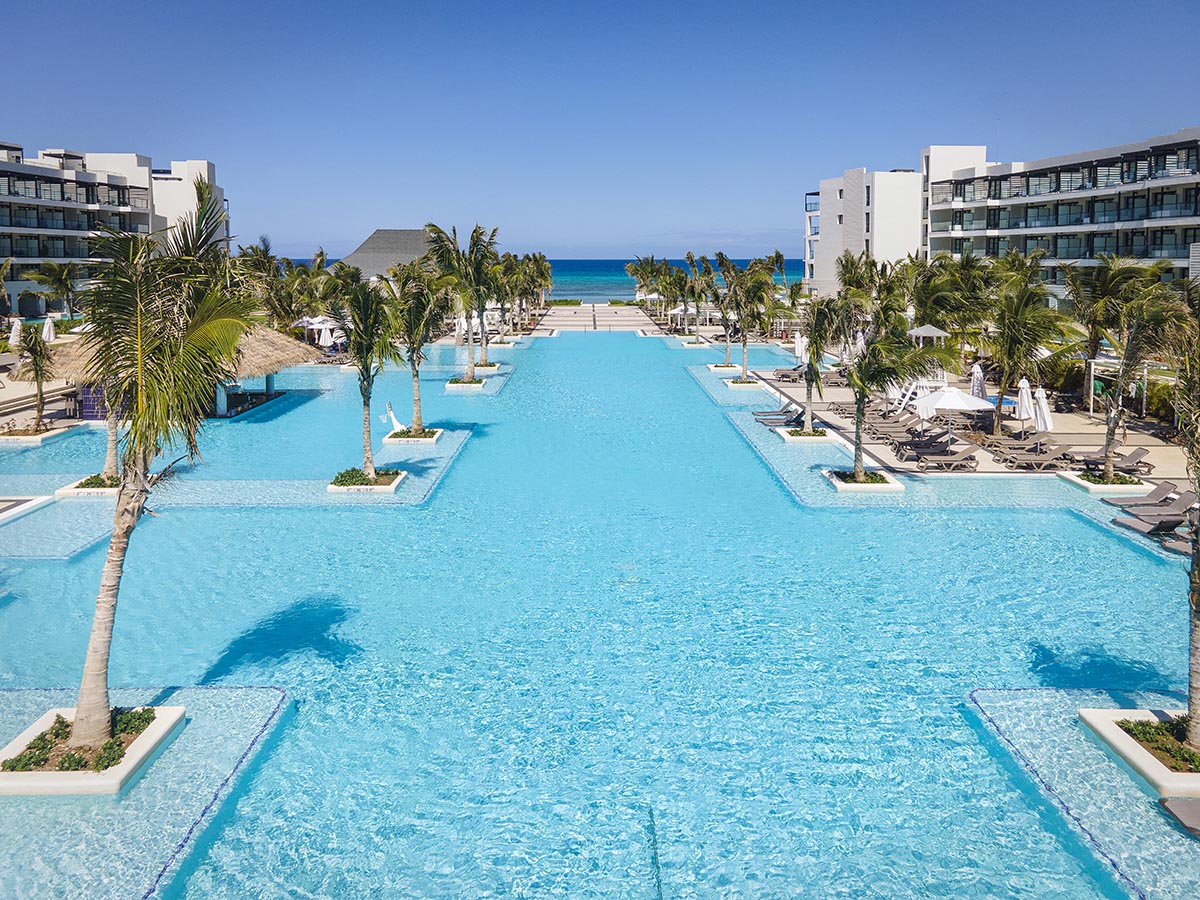 Large rectangular pool lined with palm trees, flanked by white resort buildings, with ocean and beach visible in the distance.