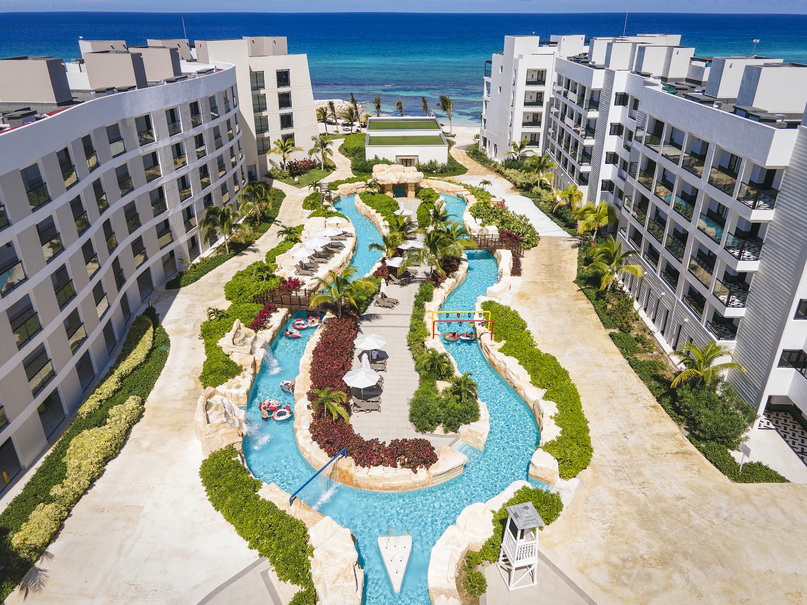 White resort buildings surround a winding pool complex with lounge chairs, tropical landscaping, and ocean view beyond.