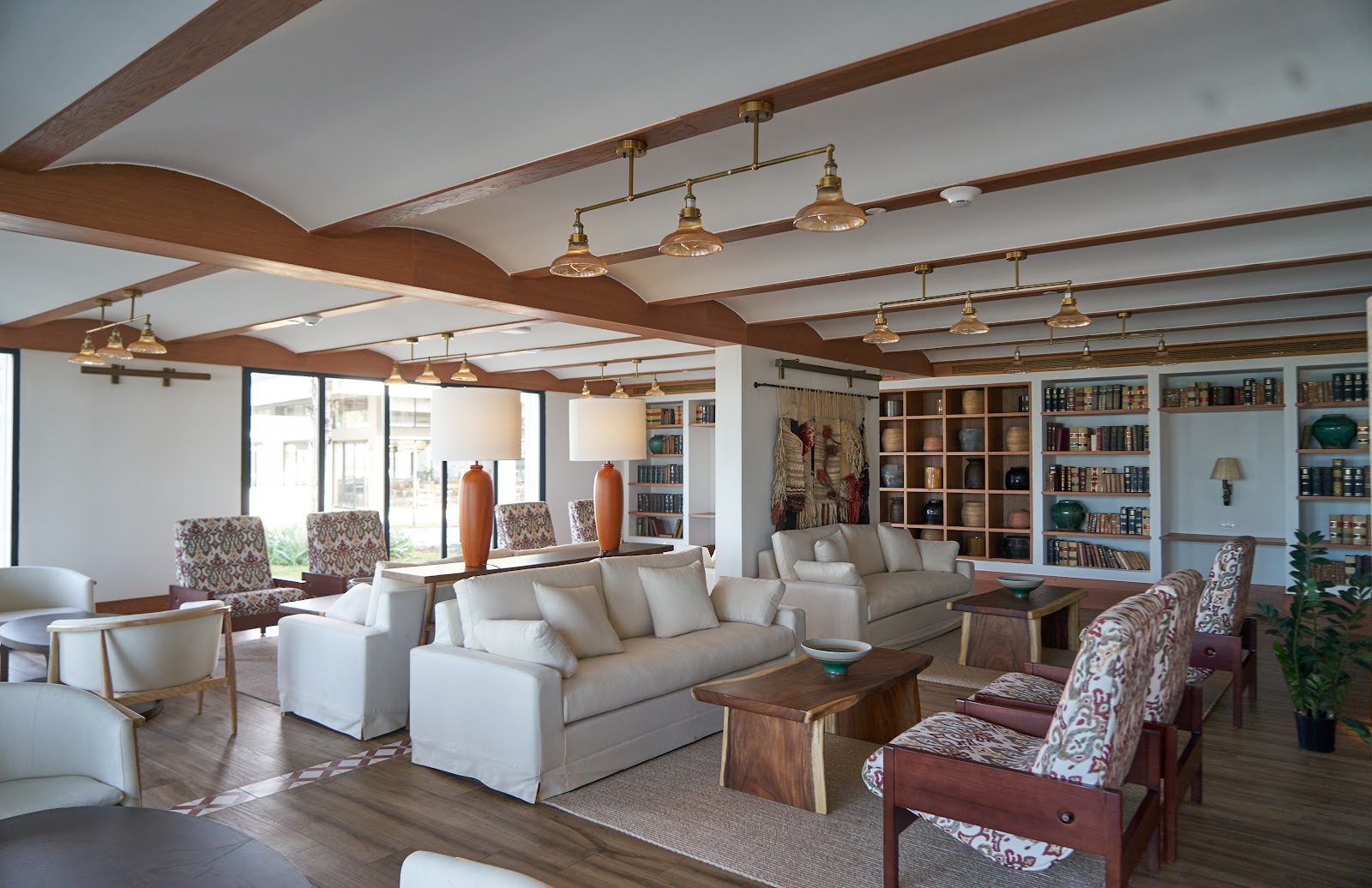 Open lounge area with exposed wood beams, cream sofas, patterned accent chairs, brass pendant lights, and built-in bookshelves at Ocean Eden Bay.