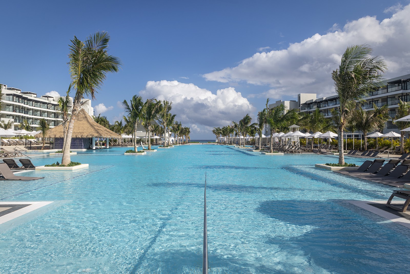 Large lagoon-style pool with turquoise water lined by palm trees, loungers, and white umbrellas, flanked by multi-story resort buildings under blue sky.