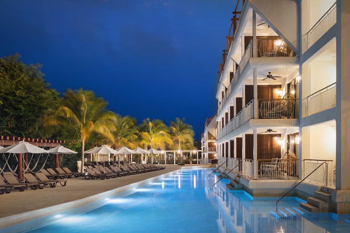 Illuminated pool runs alongside a three-story white resort building at dusk, with lounge chairs and umbrellas lining the beach beyond.