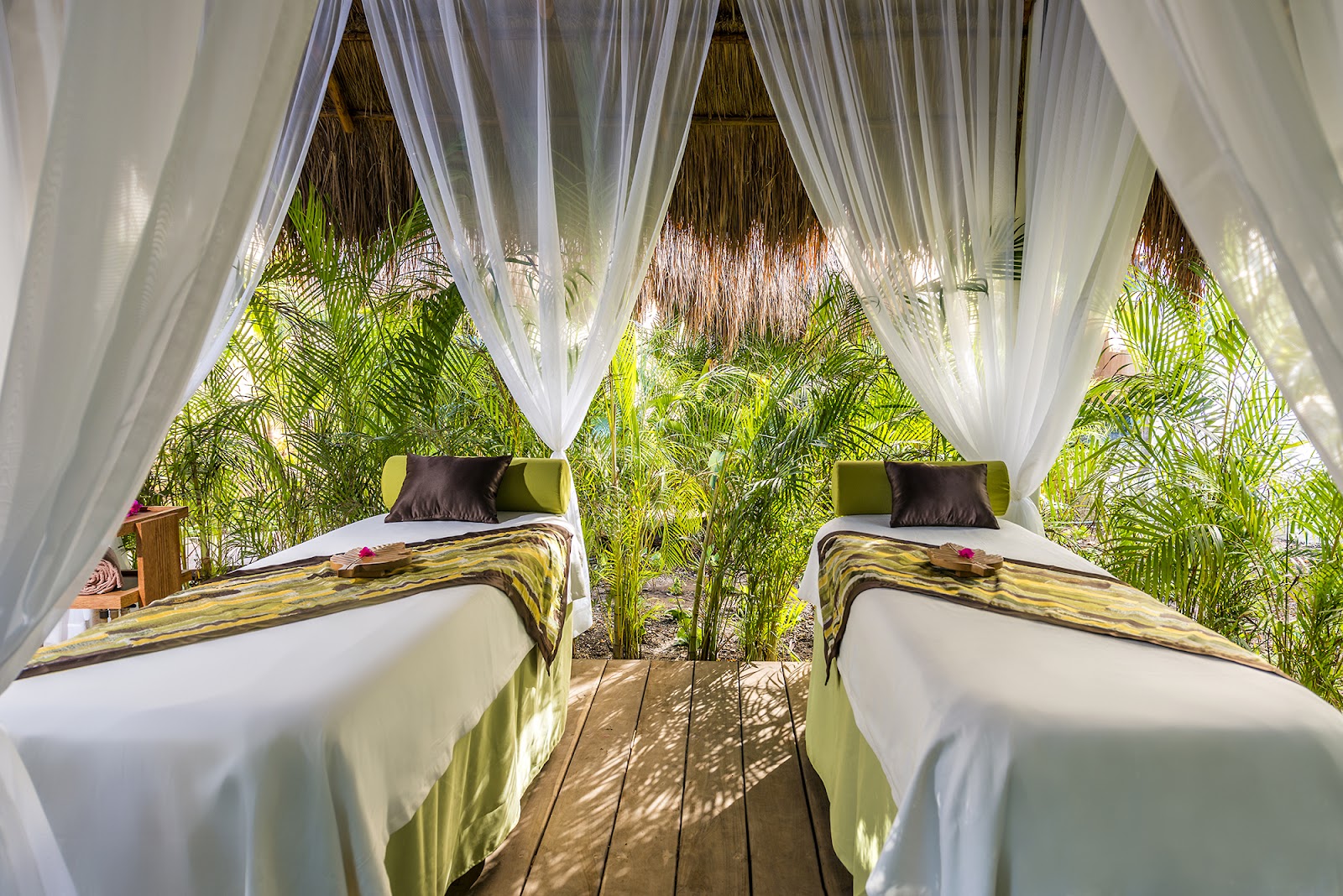 Two twin beds with white linens under flowing white curtains in a thatched-roof cabana surrounded by green palm plants.