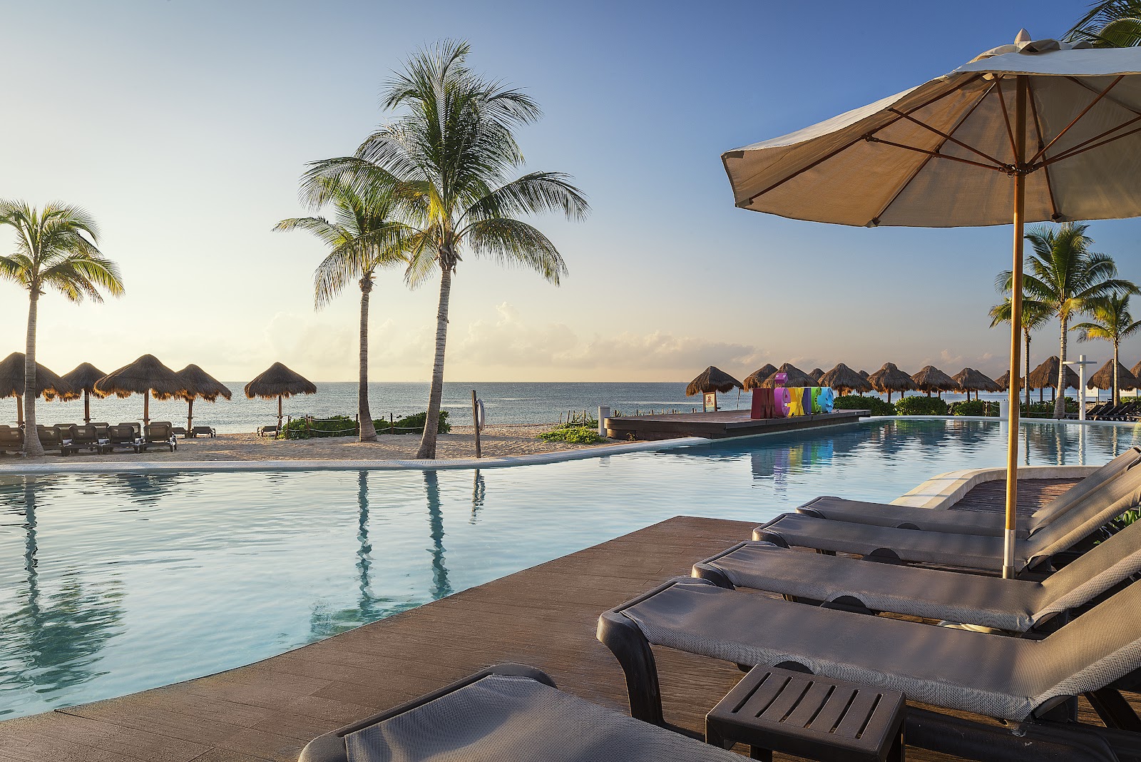 Pool deck with lounge chairs and tan umbrellas overlooking the ocean, palm trees, and thatched-roof beach palapas at Ocean Riviera Paradise.