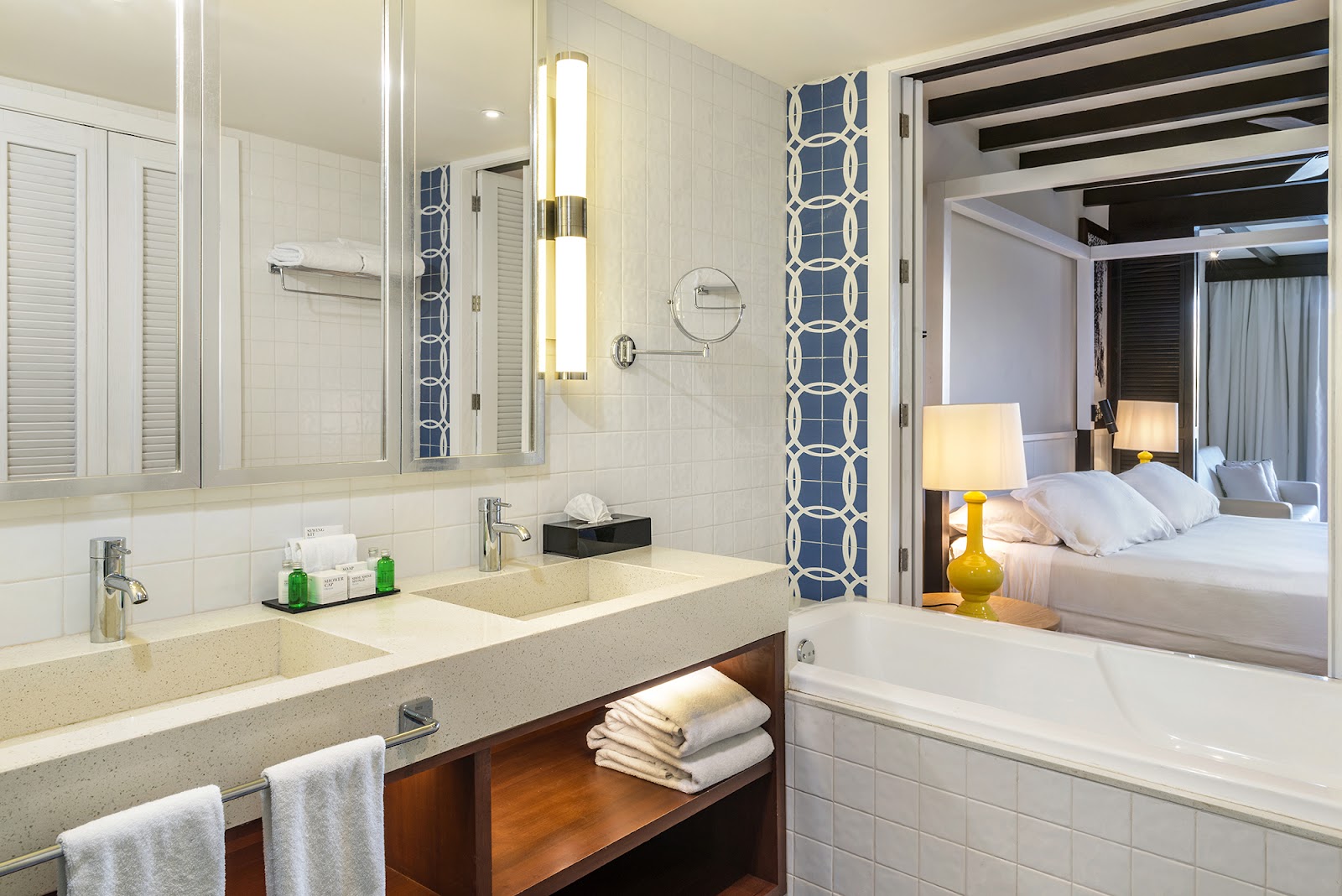 Bathroom with double vanity, glass shower, soaking tub, and open view to bedroom with yellow lamp and white bedding.