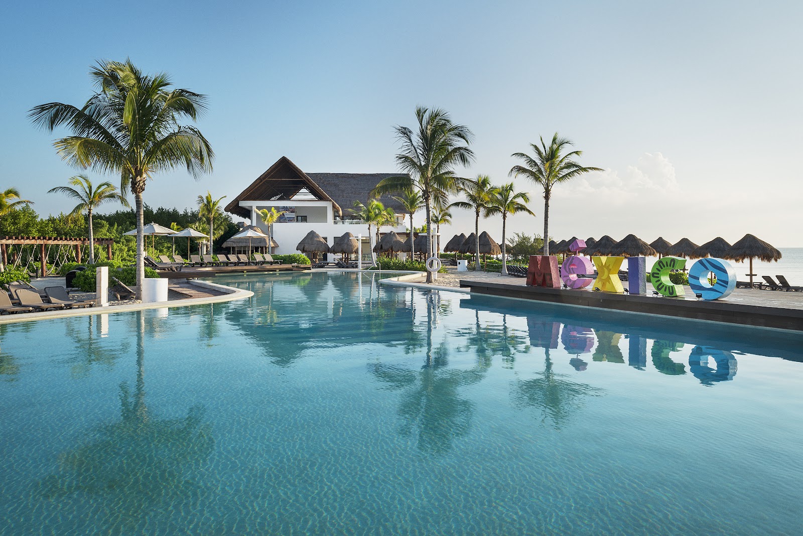 Large lagoon-style pool with colorful resort signage, thatched-roof bungalows, palm trees, and main building at Ocean Riviera Paradise.