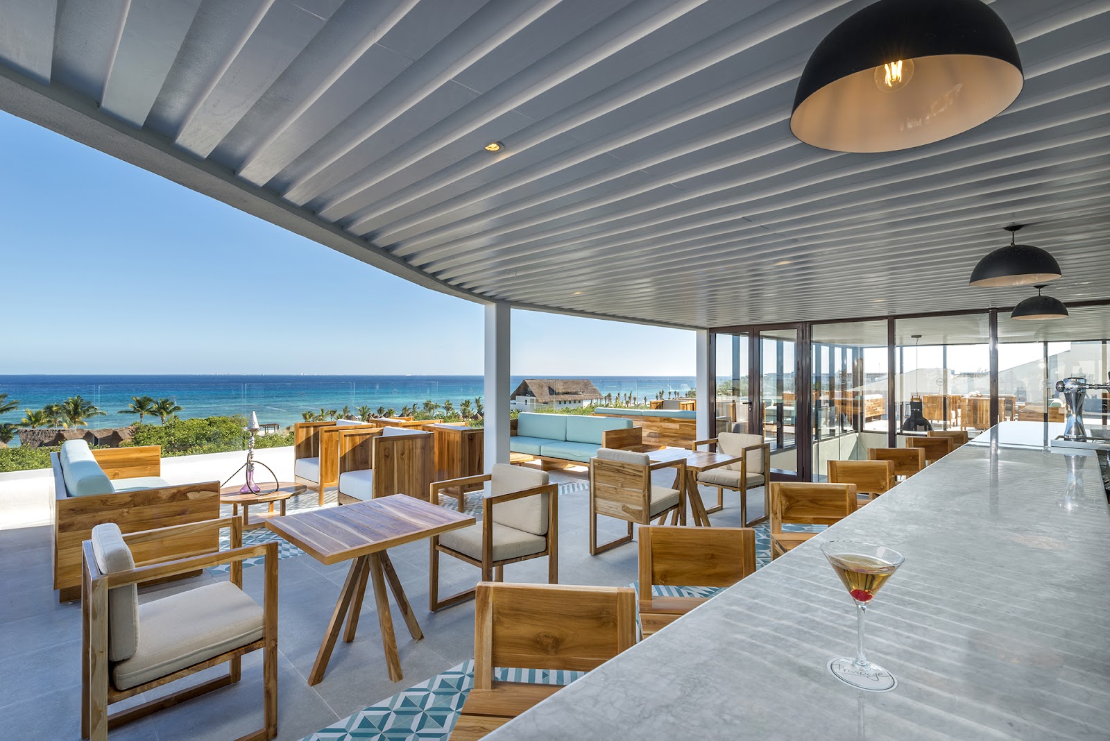 White resort buildings overlook a shallow pool with a wooden pirate ship play structure, surrounded by lounge chairs and umbrellas, with the ocean visible beyond.