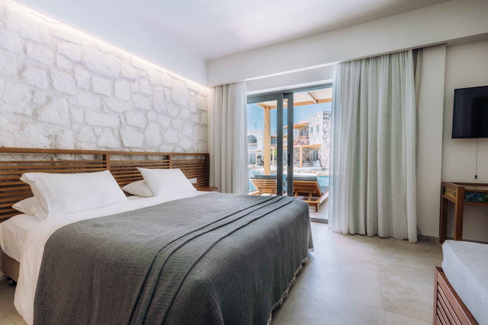 Resort bedroom with white stone wall, wood headboard bed, and glass doors opening to a sea-view balcony with wooden pergola.