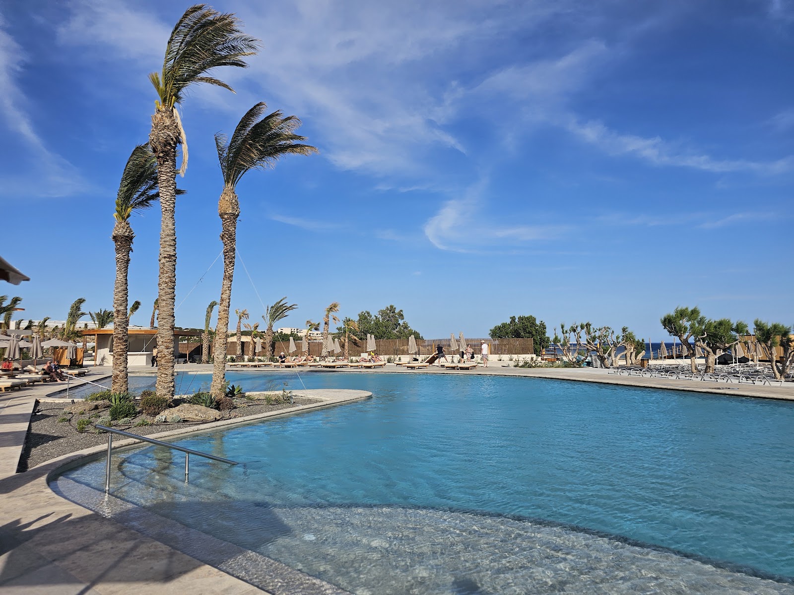 Palm trees sway above a turquoise lagoon pool at Ostria Resort & Spa, with beach loungers and tan buildings visible along the waterfront.