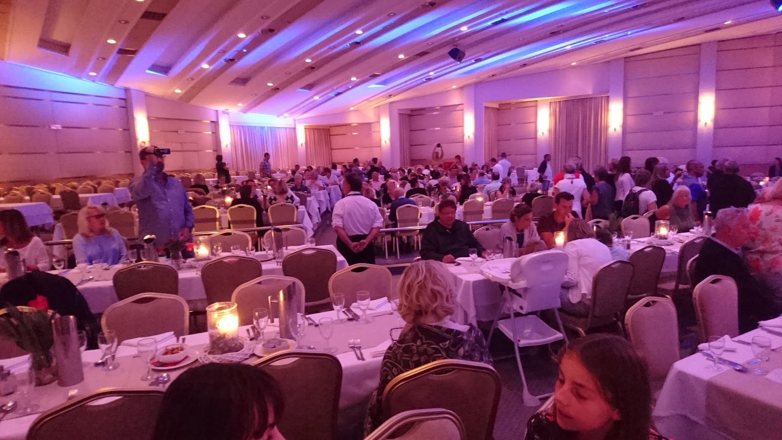 Ballroom dining hall with purple and blue ambient lighting, white-clothed tables, guests seated throughout, and glowing centerpieces.