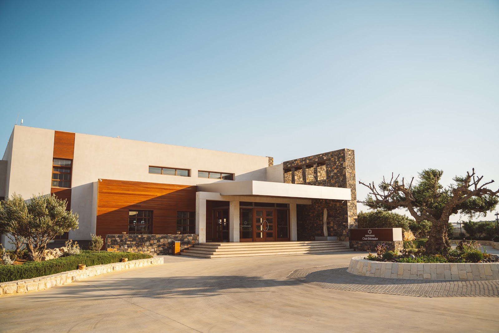 Modern entrance of Ostria Resort & Spa with cream and stone facade, wooden accents, and manicured landscape under clear sky.