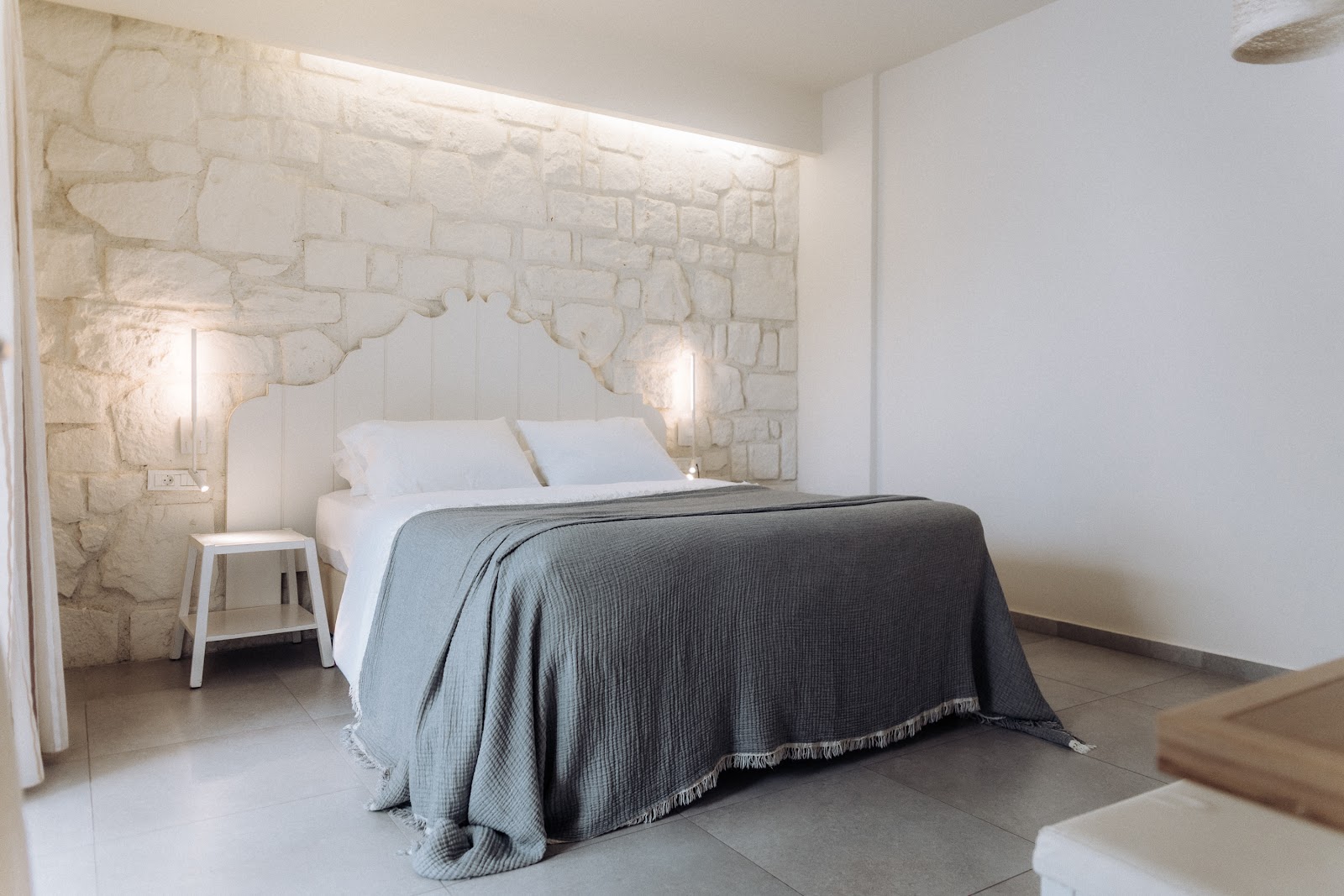 Modern resort bedroom with a queen bed draped in a gray throw, white pillows, cream stone accent wall with map design, and bedside lighting.