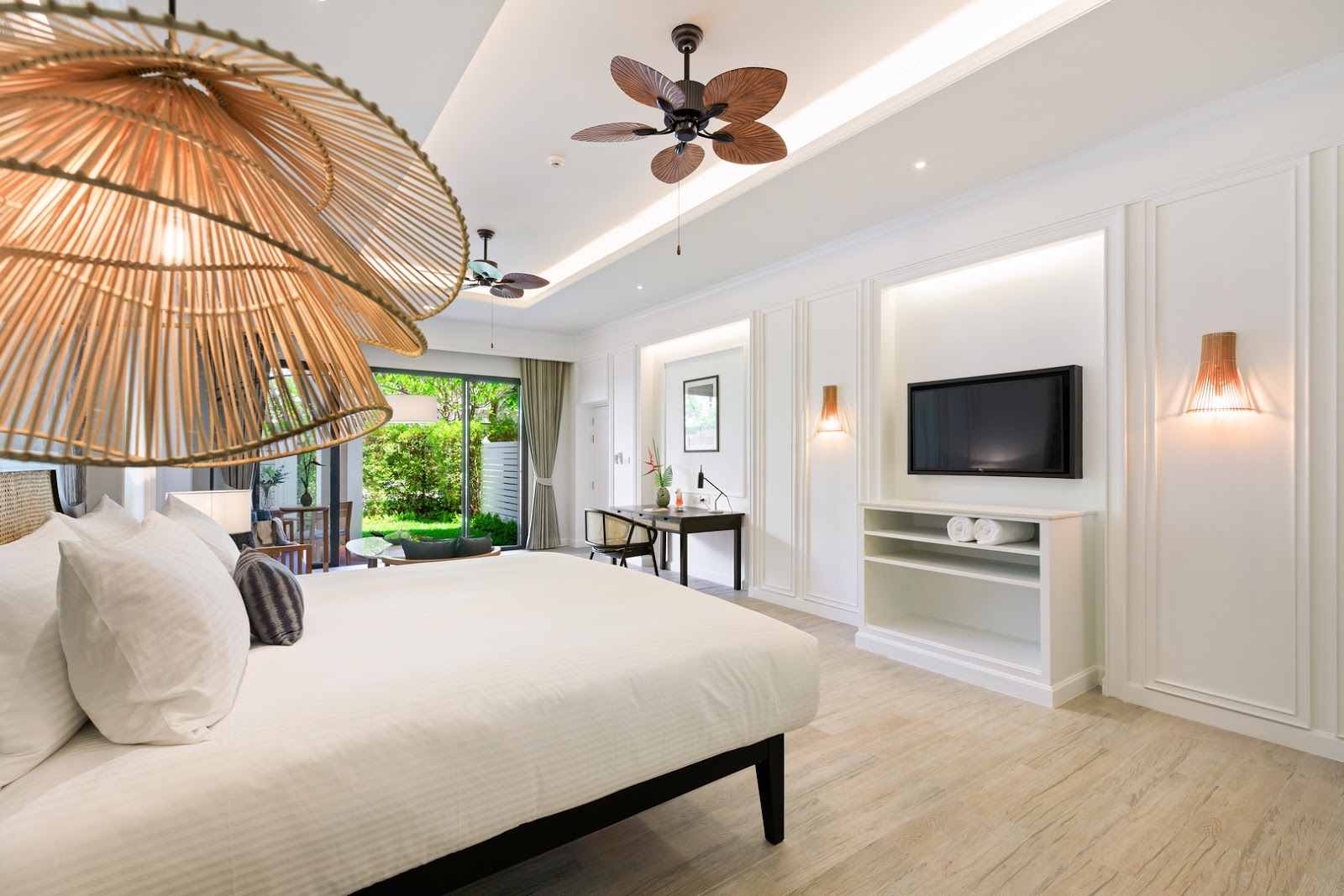 Spacious resort bedroom with woven pendant lights, white bedding, wall-mounted TV, and glass doors opening to tropical grounds.