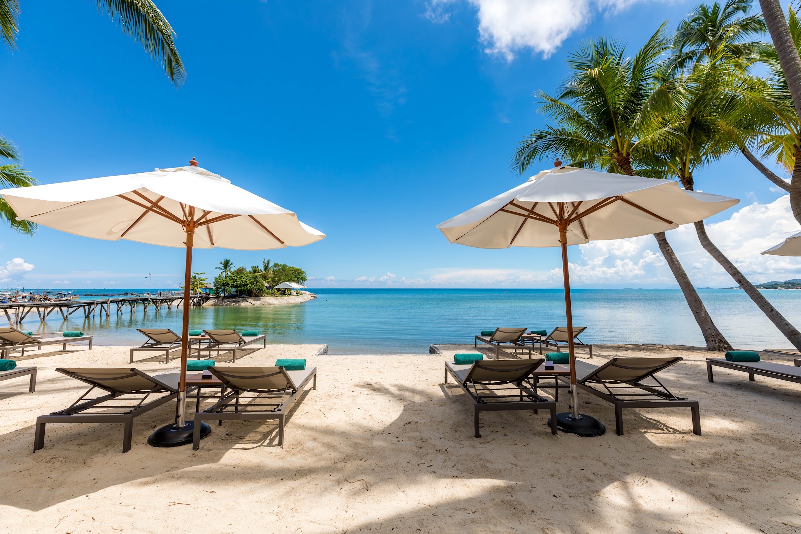 Beach with lounge chairs and cream umbrellas facing turquoise water, palm trees, and a wooden pier at OUTRIGGER Koh Samui Beach Resort.