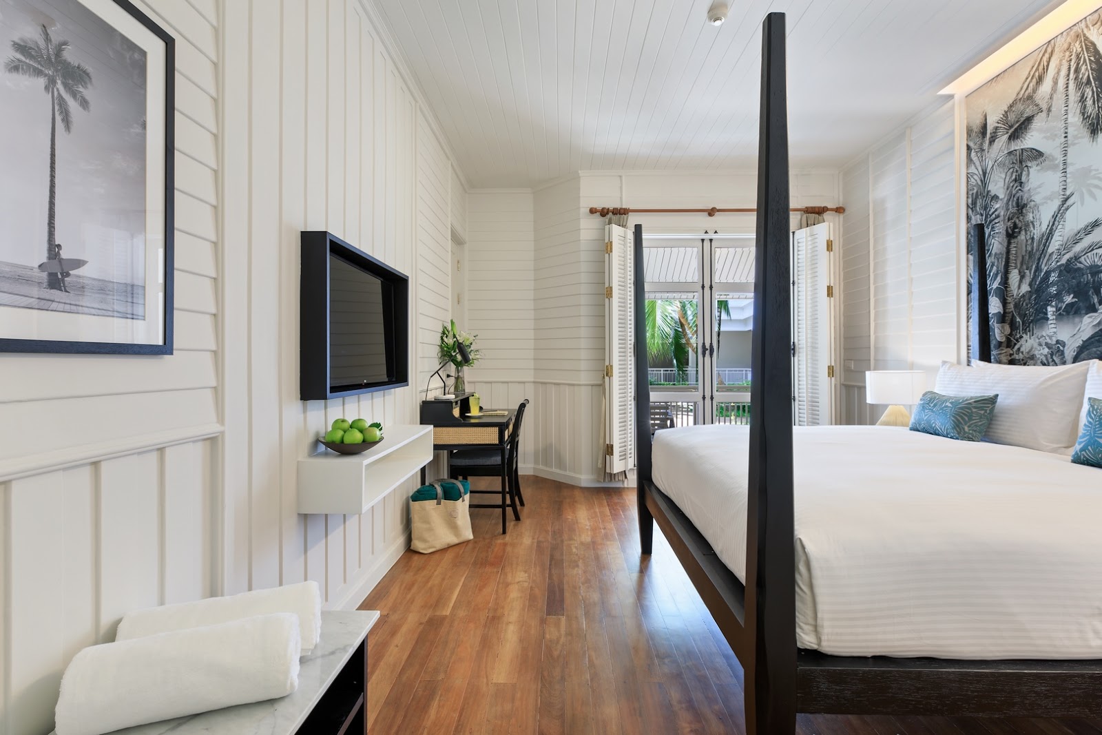 Resort guest room with black four-poster bed, white shiplap walls, wooden floors, TV, desk area, and glass doors opening to a balcony with ocean view.