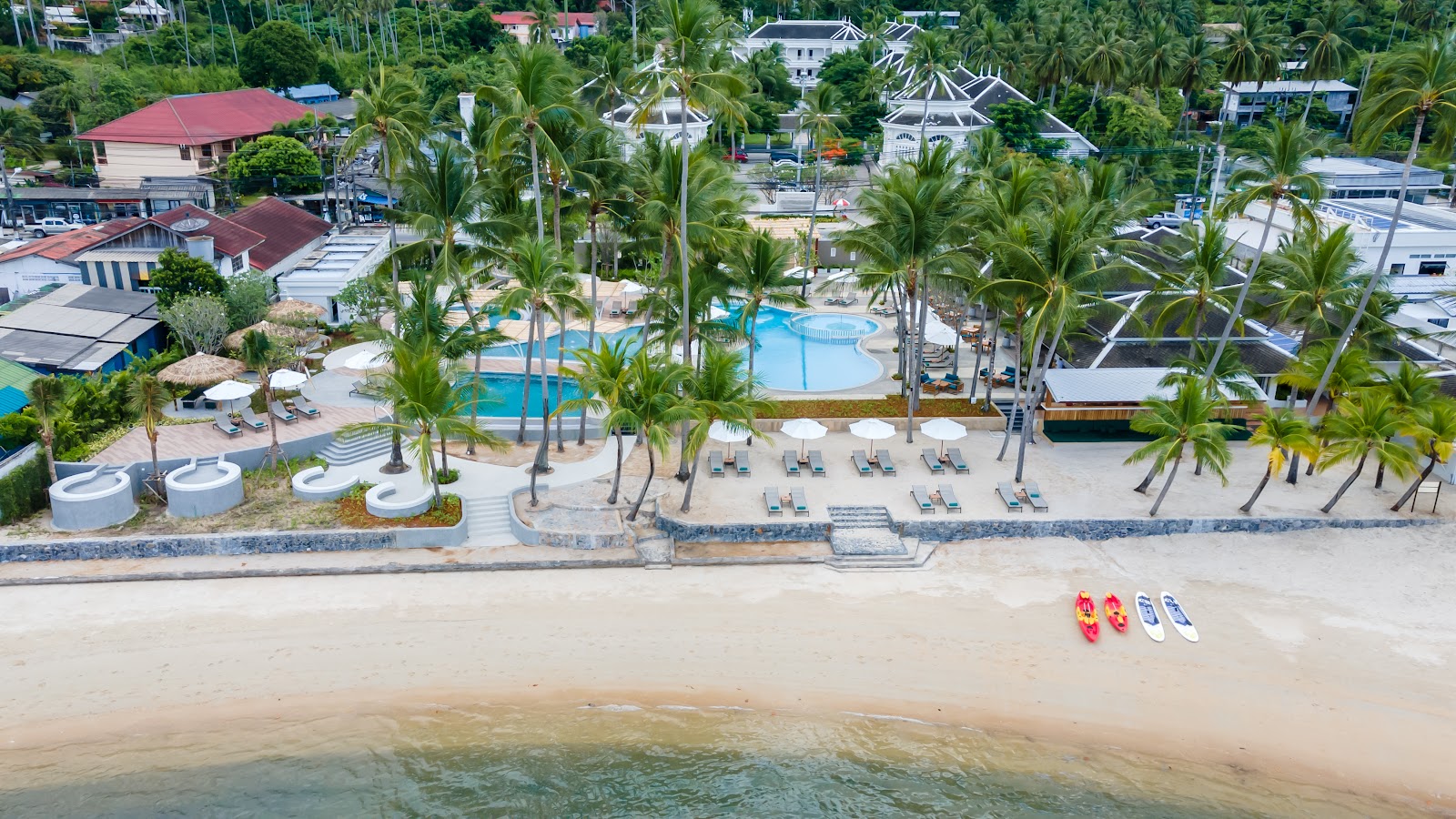 Sandy beach with turquoise water, kayaks on shore, and forested mountains rising behind beachfront buildings and palm trees.