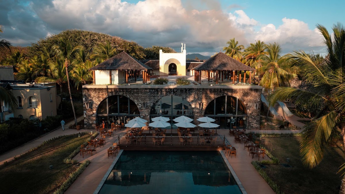 Pool deck with white umbrellas and wooden chairs facing a stone archway building, palm trees and thatched-roof structures surrounding the OUTRIGGER Mauritius Beach Resort.