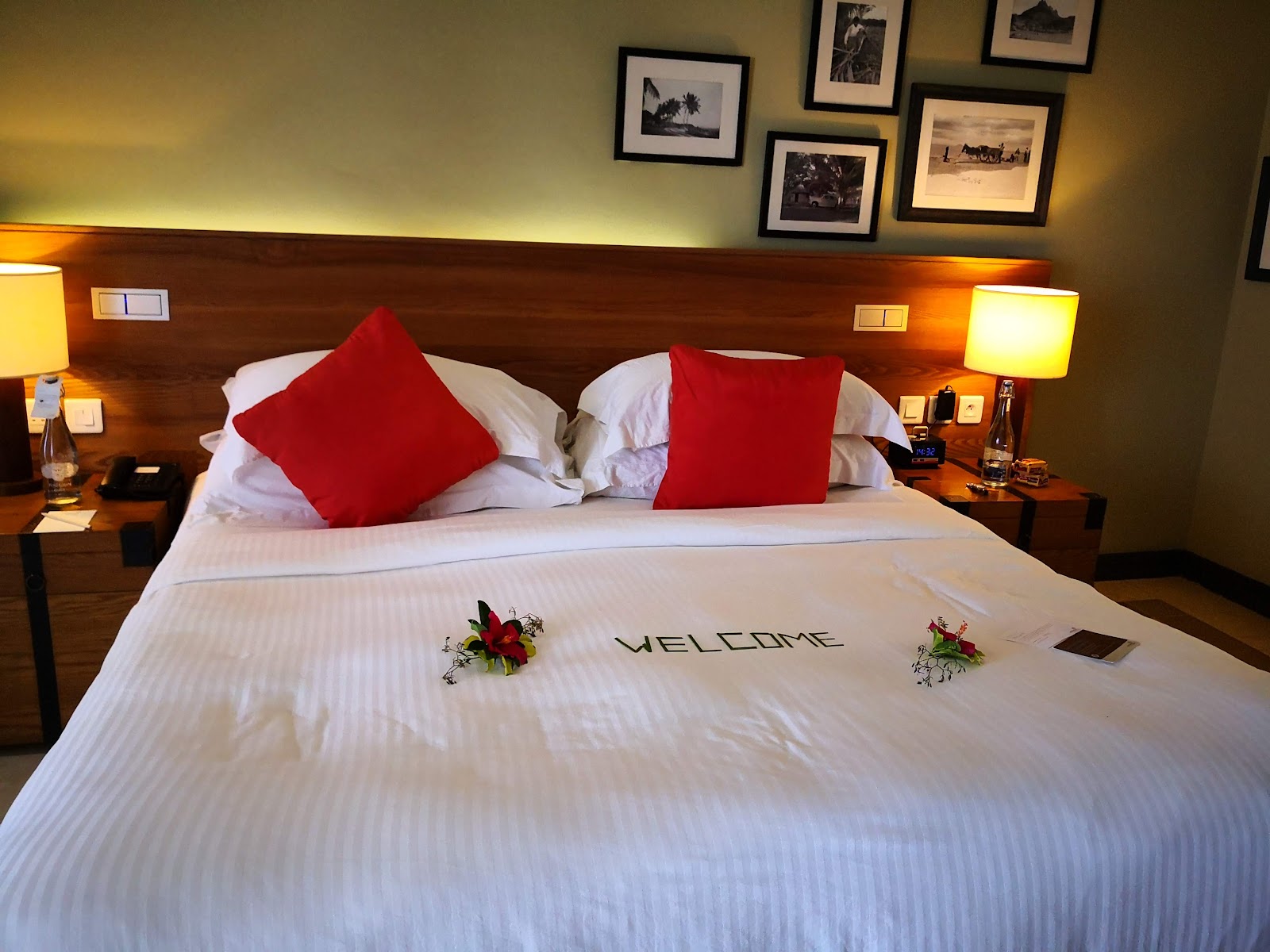 Resort bedroom with a queen bed dressed in white linens, red accent pillows, and a welcome message, flanked by wood nightstands with warm lighting.