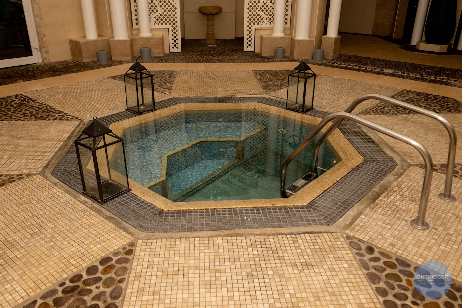 Indoor thermal bath with turquoise water, tiled floor, metal railings, and decorative lanterns in a spa area.