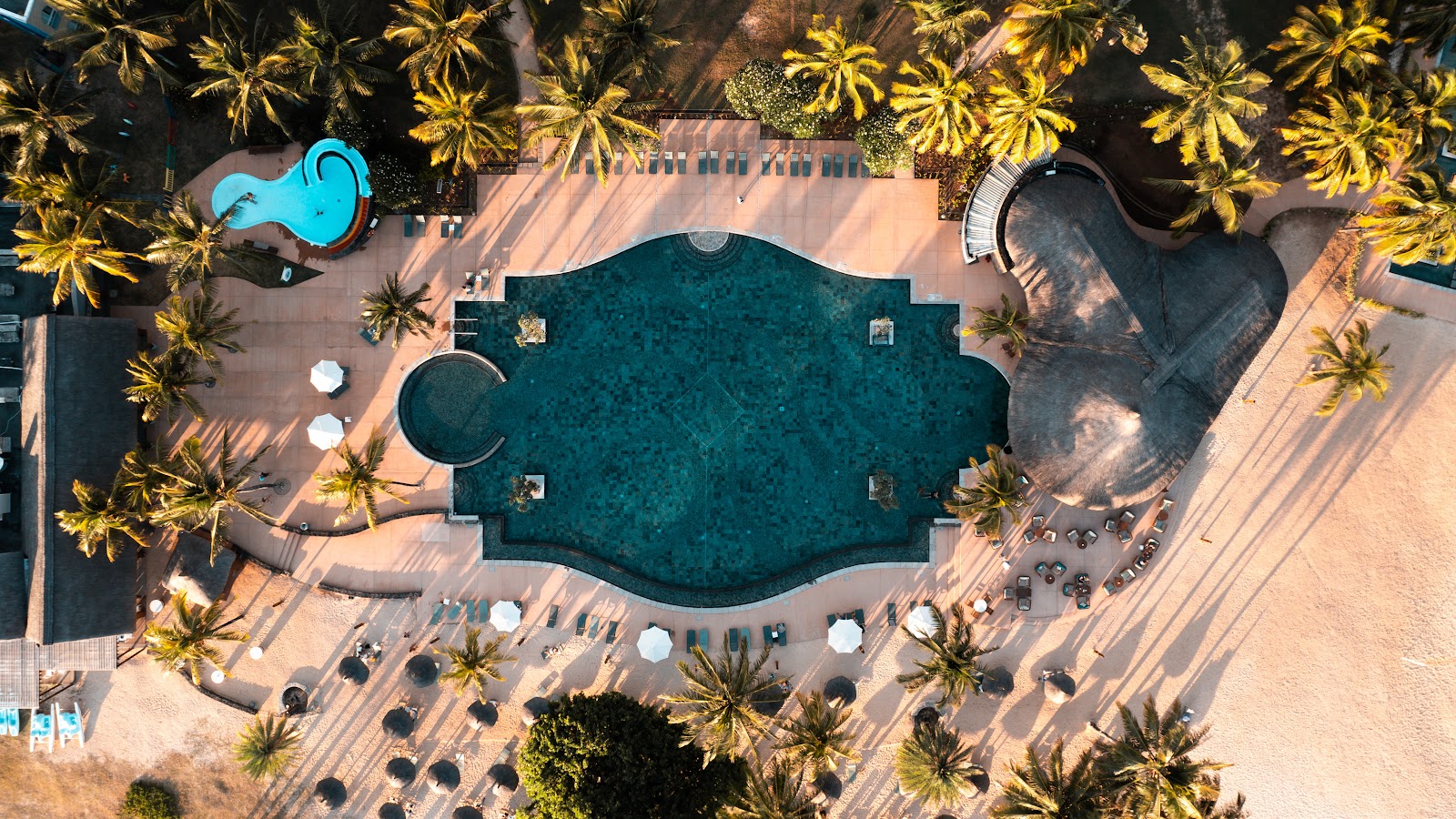 Overhead view of an ornate turquoise pool surrounded by sandy deck, umbrellas, lounge chairs, palm trees, and resort buildings at OUTRIGGER Mauritius Beach Resort.