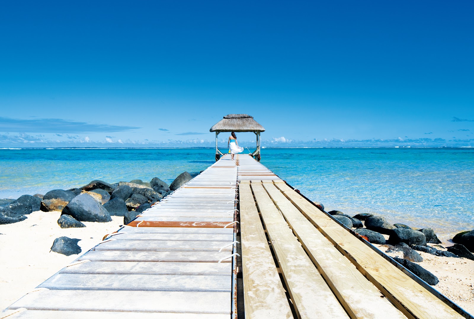 Woman in white dress sits in thatched gazebo at end of wooden pier extending into turquoise lagoon at OUTRIGGER Mauritius Beach Resort.