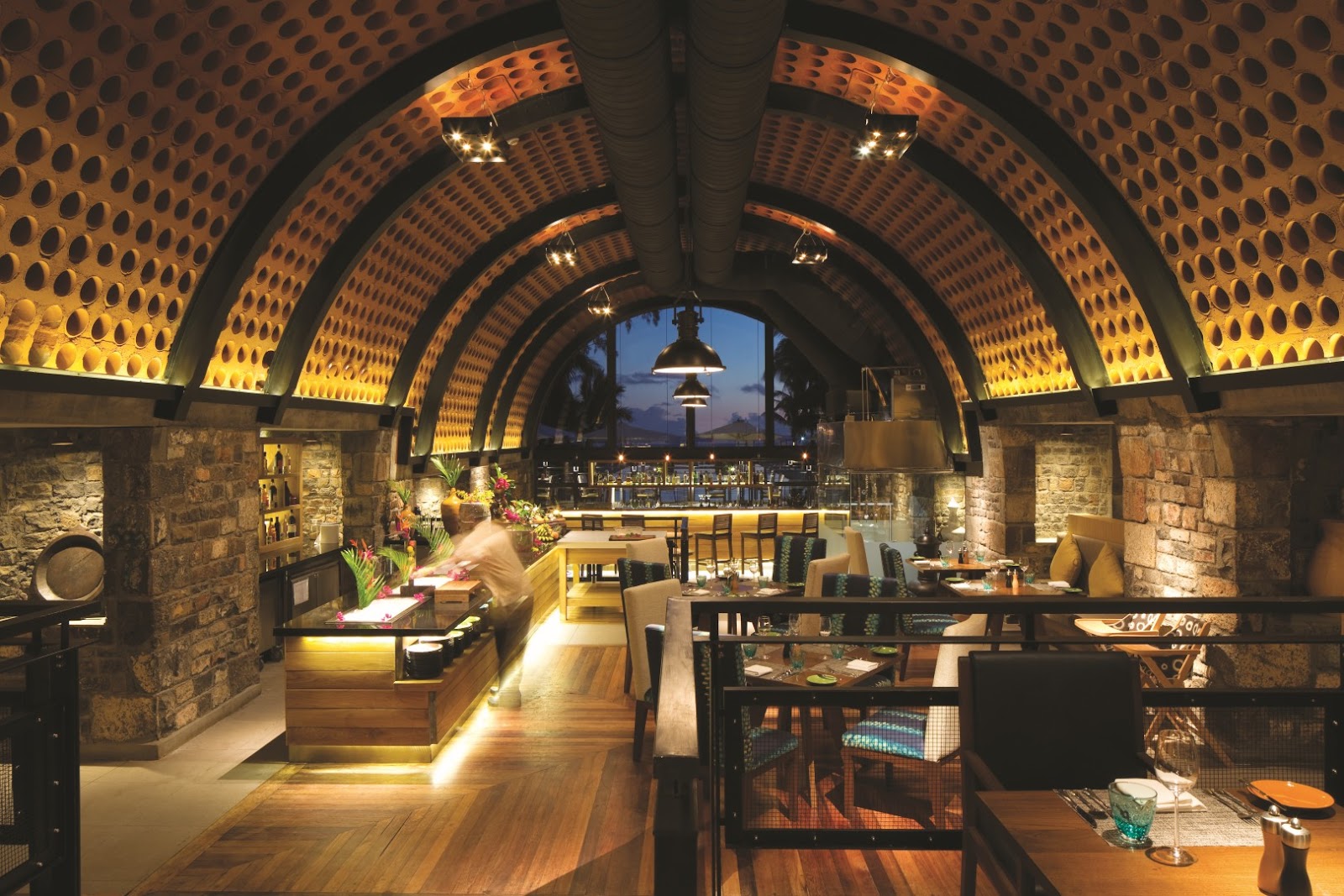 Arched restaurant interior with perforated stone ceiling, wooden floors, and dining tables overlooking tropical ocean at dusk.