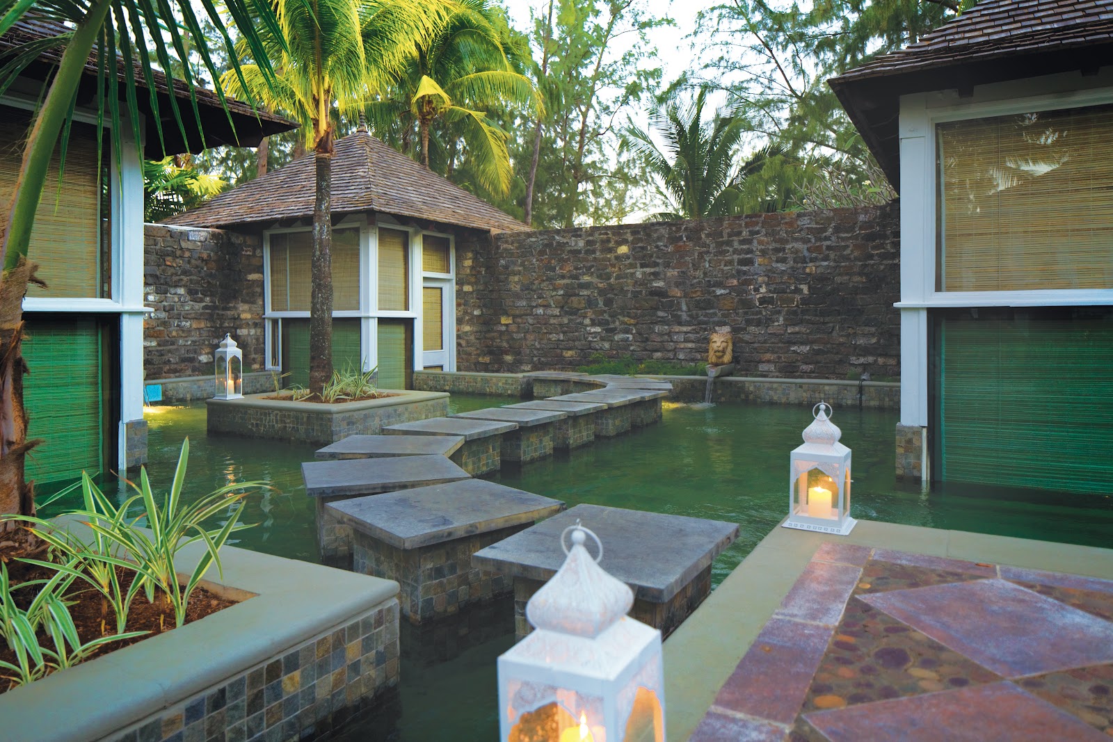 Stone villa courtyard with stepping stones across a reflecting pool, palm trees, white lanterns, and green shuttered doors.