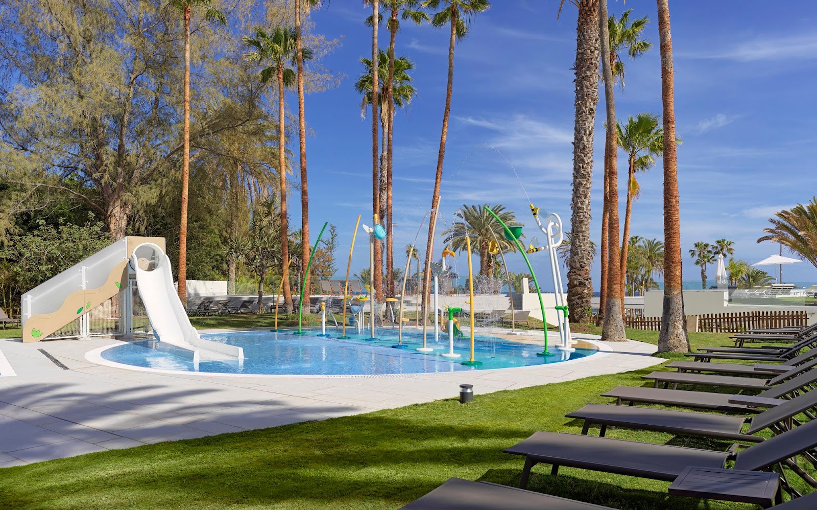 Children's splash pool with white slide and water jets surrounded by tall palms, loungers, and ocean view at Paradisus Gran Canaria.