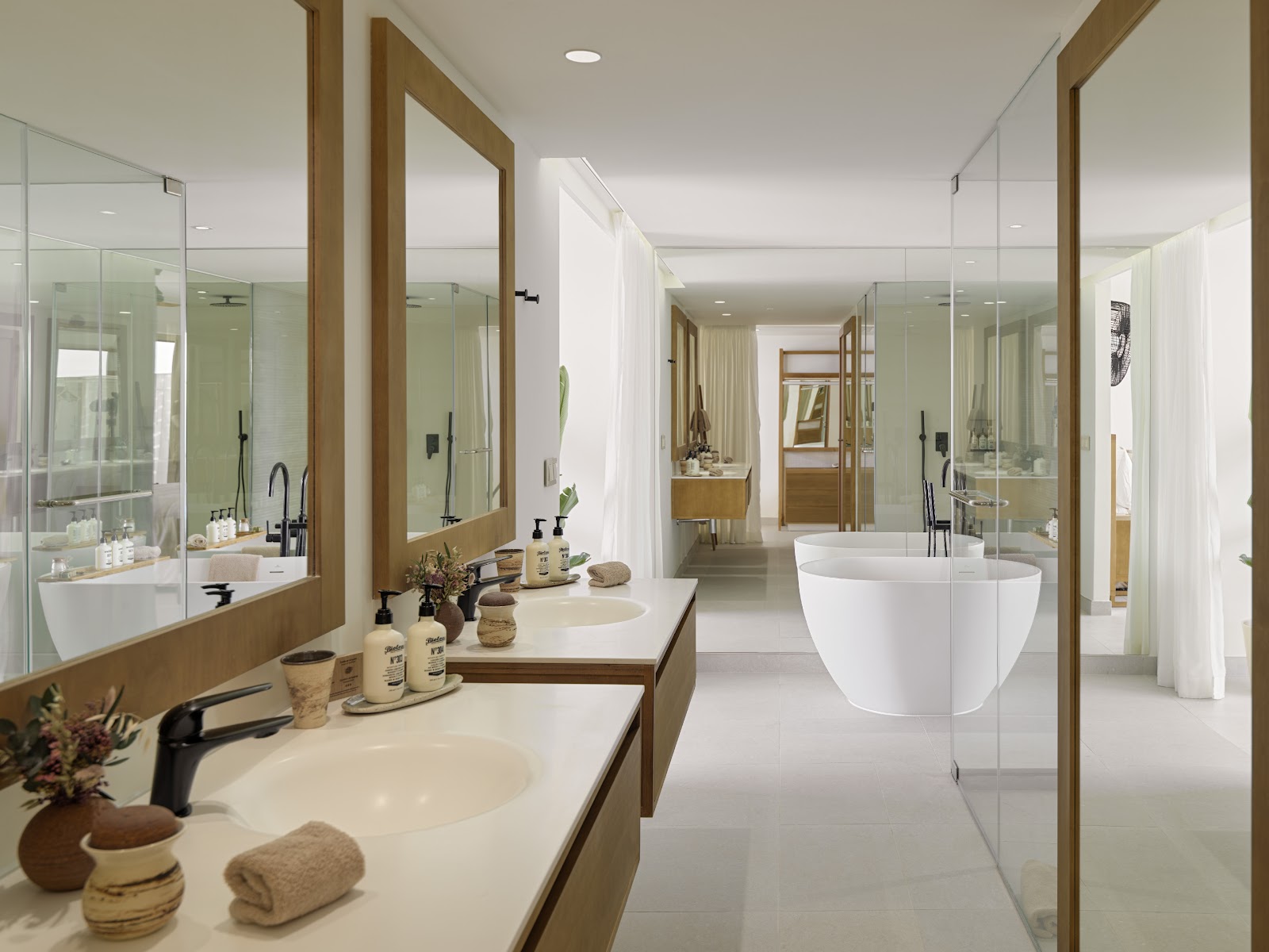 Modern resort bathroom with dual vanities, large mirrors with wood frames, a freestanding white tub, and glass-enclosed shower areas.