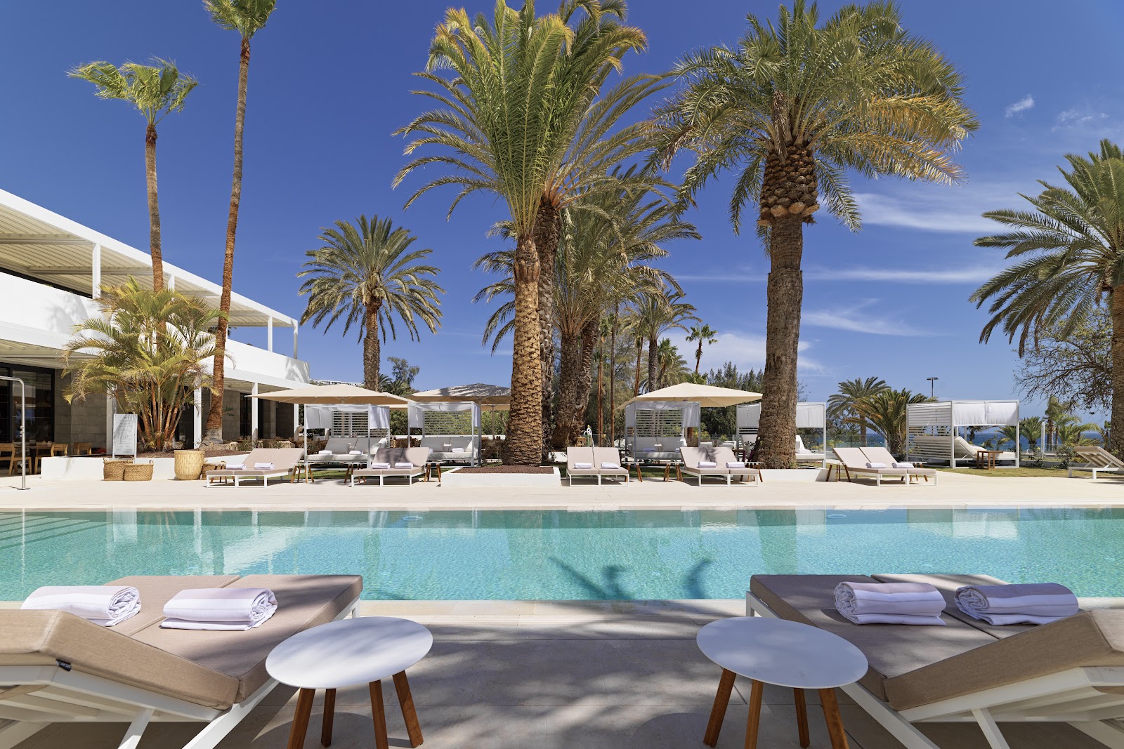 Clear turquoise pool with palm tree shadows, white loungers, and tall resort building under blue sky at Paradisus Gran Canaria.