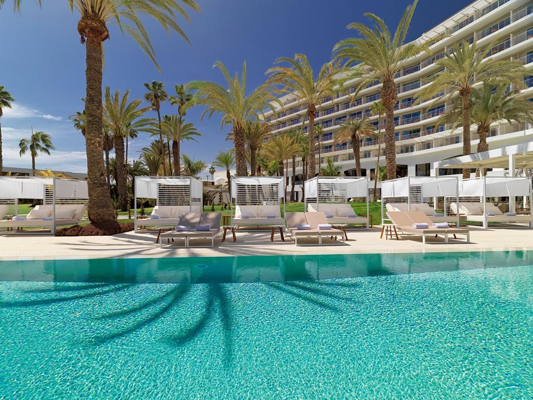 Modern lounge with cream seating, wood shelving, and large windows overlooking palm trees and pool at Paradisus Gran Canaria.